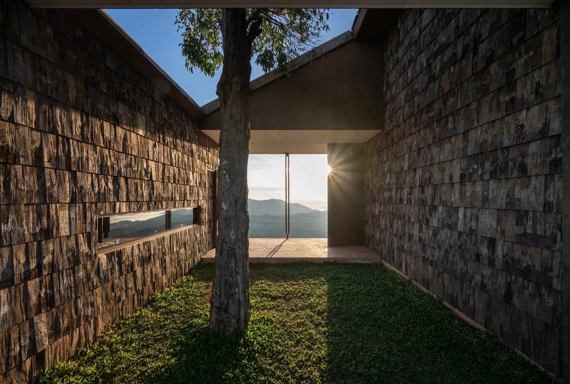 Grassy courtyard flanked by timber-shingled walls with sunlight streaming through a corridor to mountain views