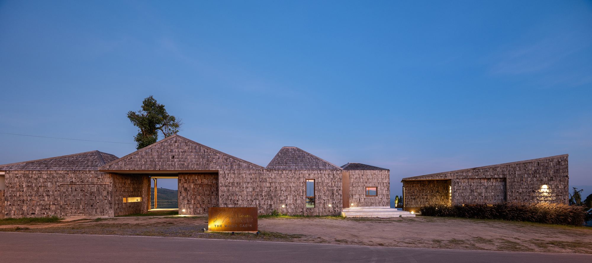 Evening view of shingled pavilions with illuminated openings arranged along a gravel clearing at twilight