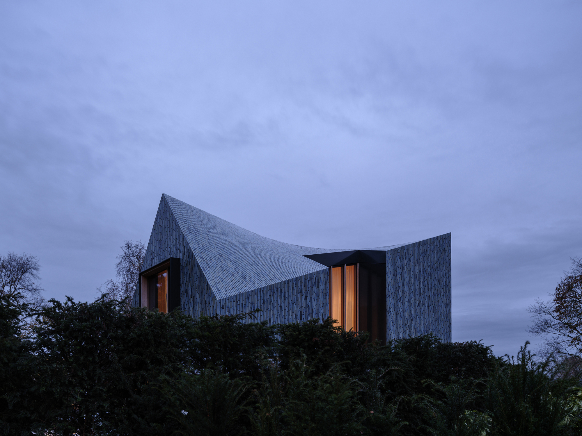 Illuminated windows glowing in the ceramic-clad volume above hedges at dusk