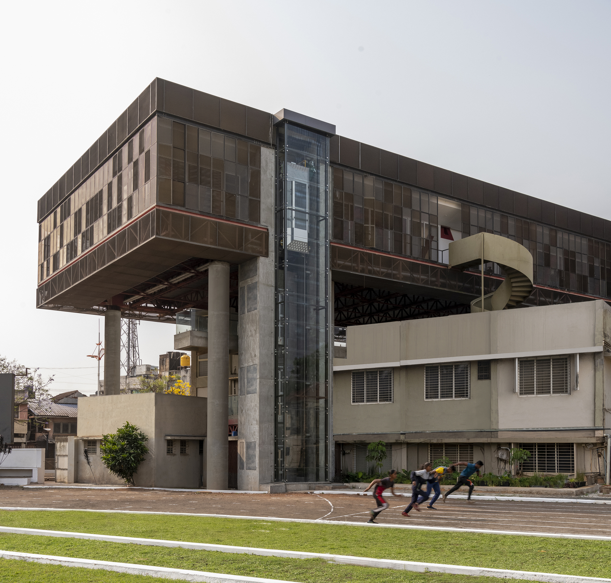 Glazed elevator shaft rising between exposed structural trusses as athletes sprint across the adjacent track