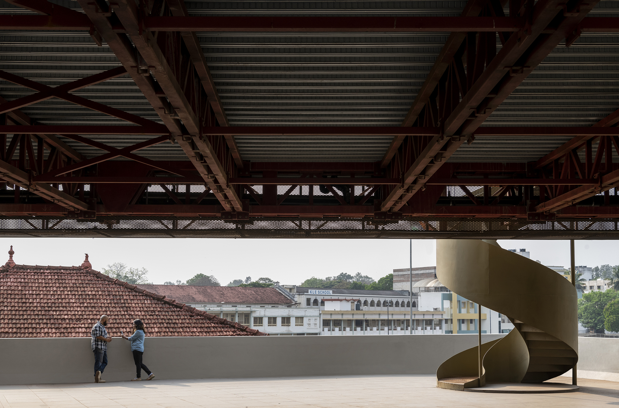 Rooftop terrace with a helical metal staircase and two figures walking past exposed trusses overhead
