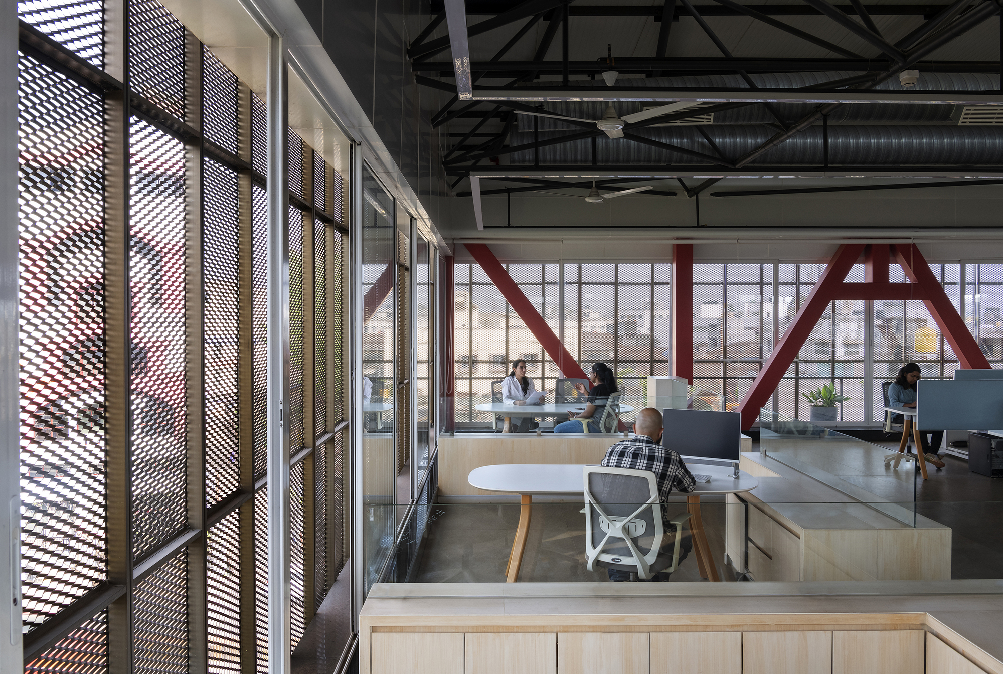 Open office space with exposed red structural trusses and perforated screen walls with workers at desks