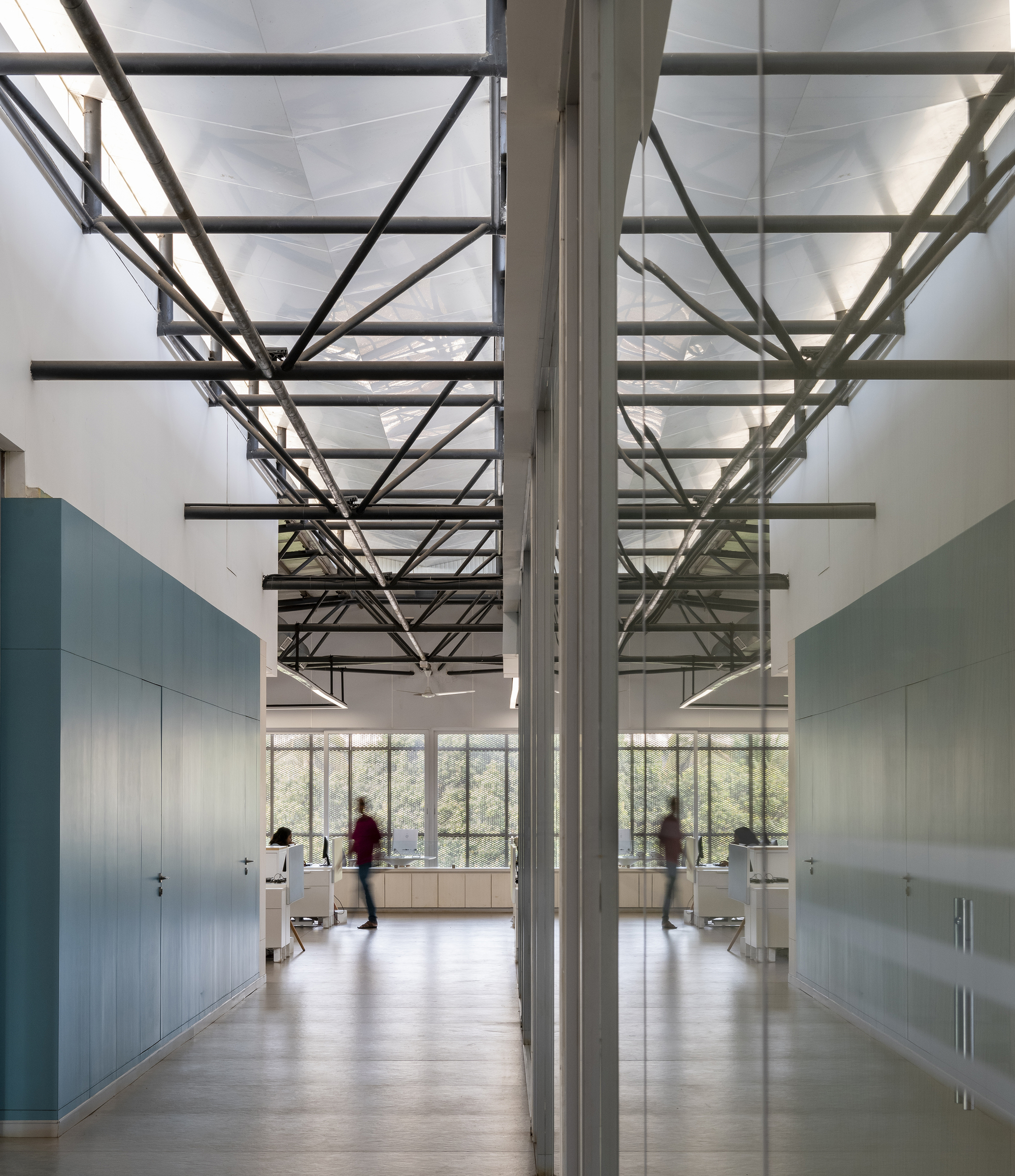Double-height corridor with exposed steel roof trusses and mirrored wall reflecting people walking through