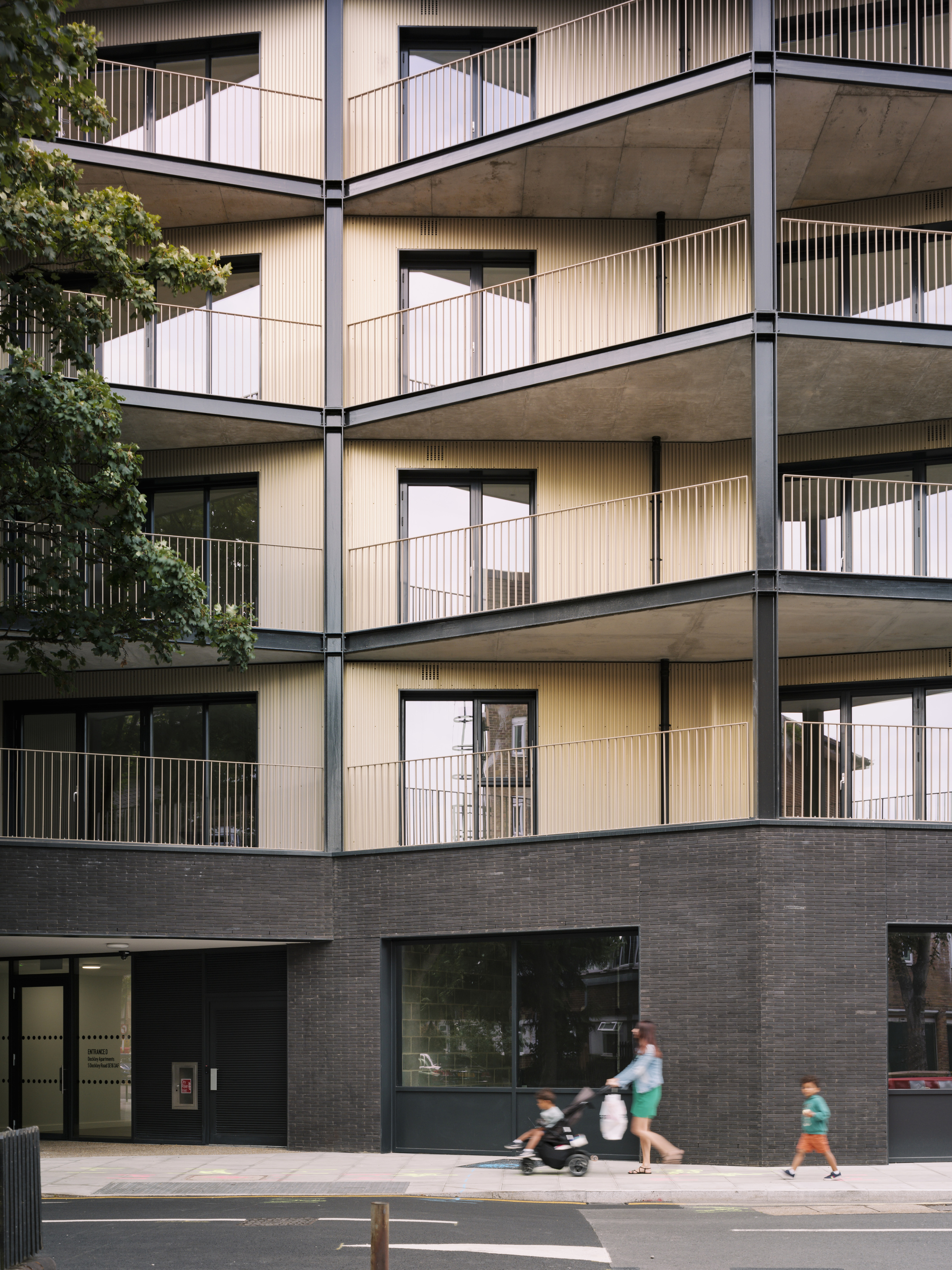 Stacked balconies with vertical metal screening as pedestrians and cyclist move along the pavement below