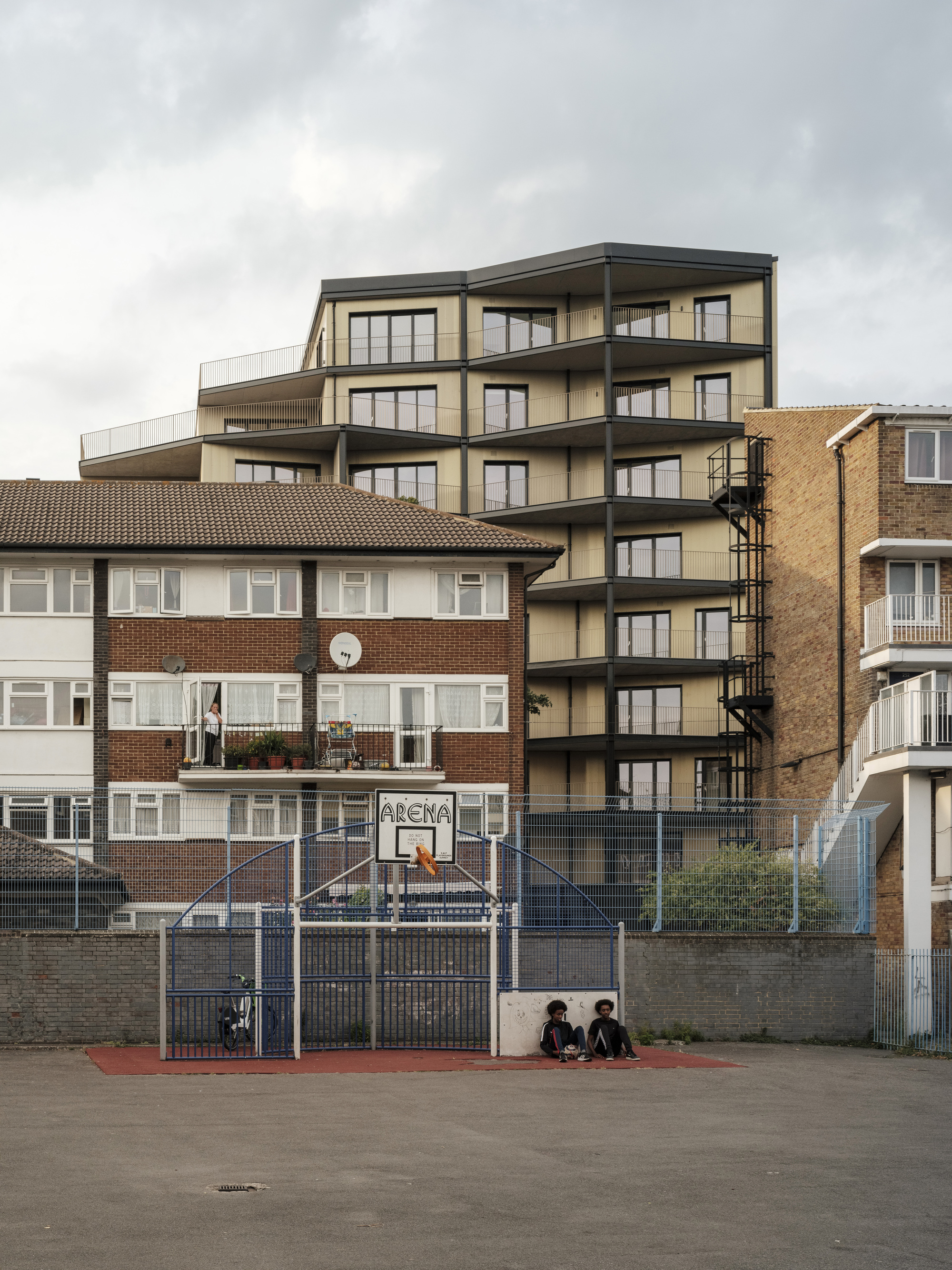 View of the residential building rising behind a brick housing block and basketball court with two seated figures