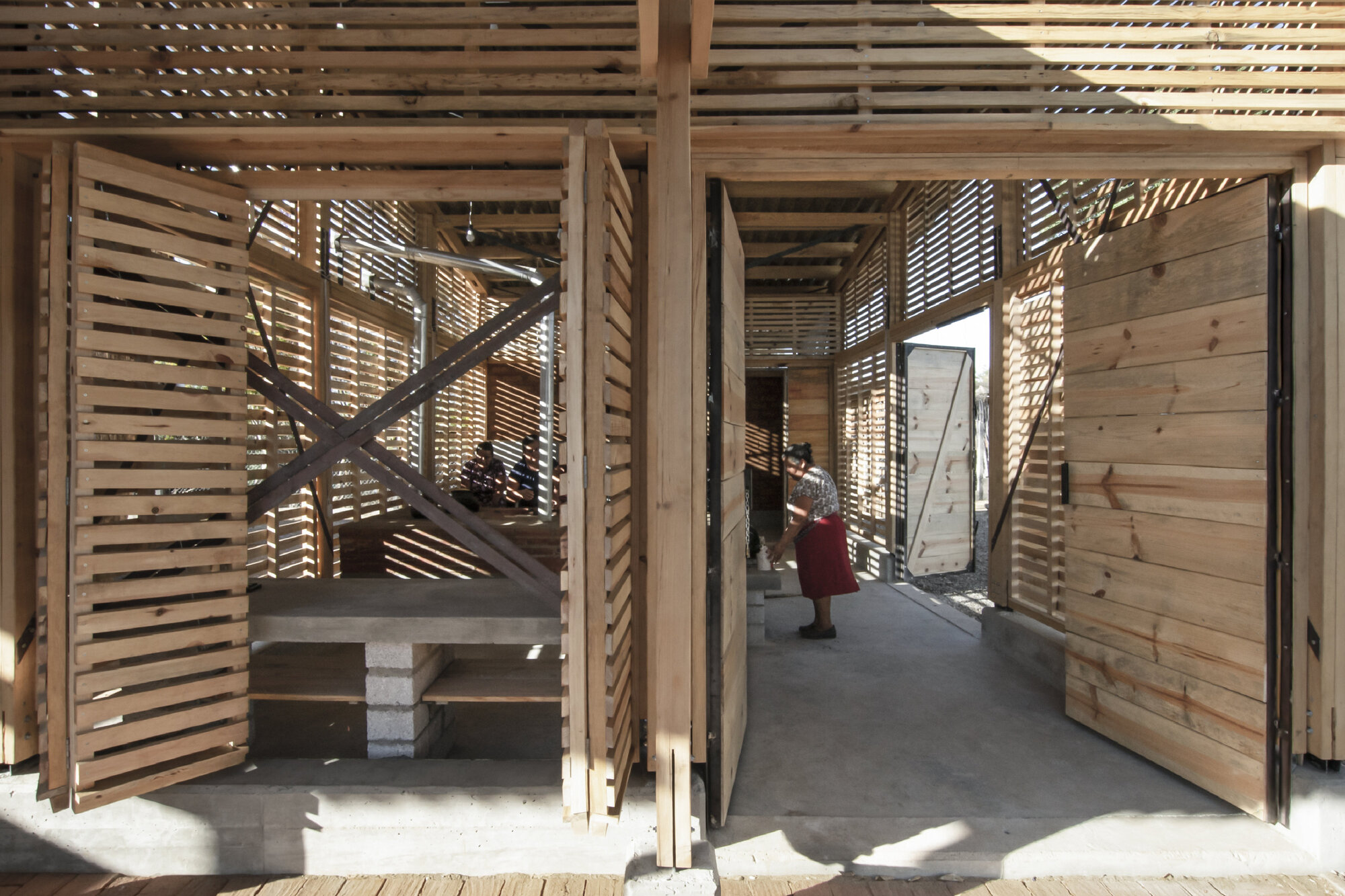Interior threshold with diagonal timber bracing and slatted walls filtering dappled sunlight