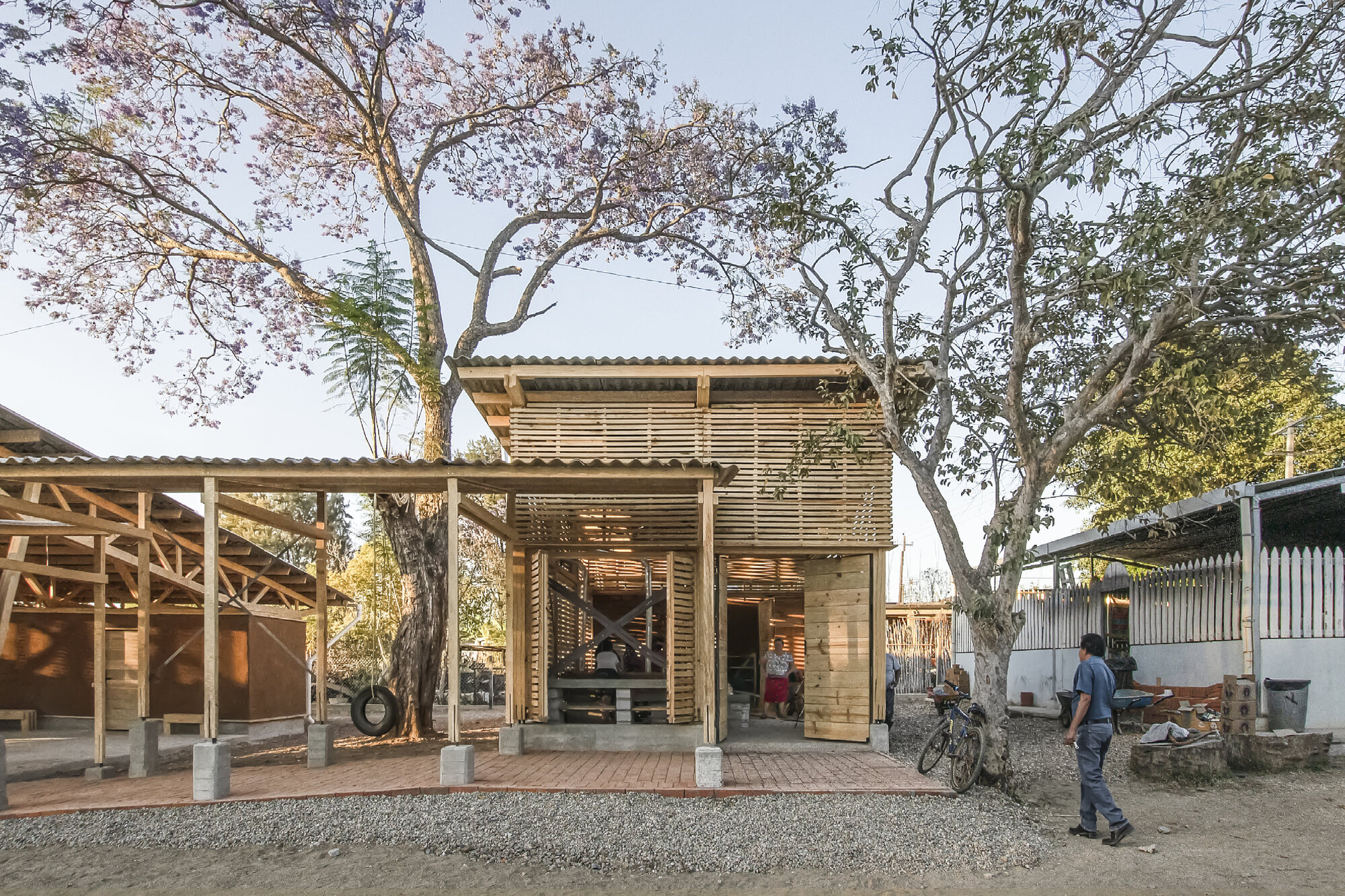 Two-story timber pavilion with louvered screens and concrete base beneath mature trees at dusk