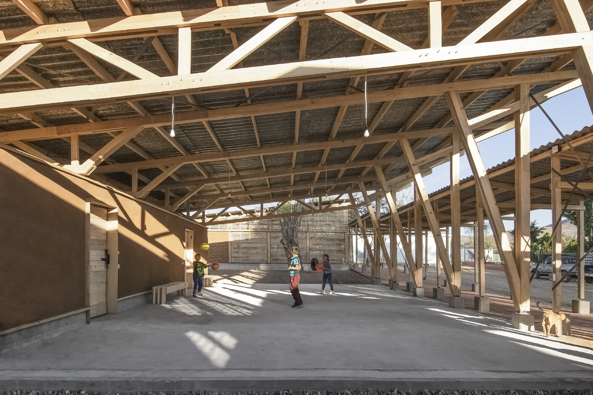 Interior of open pavilion showing timber truss structure with corrugated ceiling and children on concrete floor