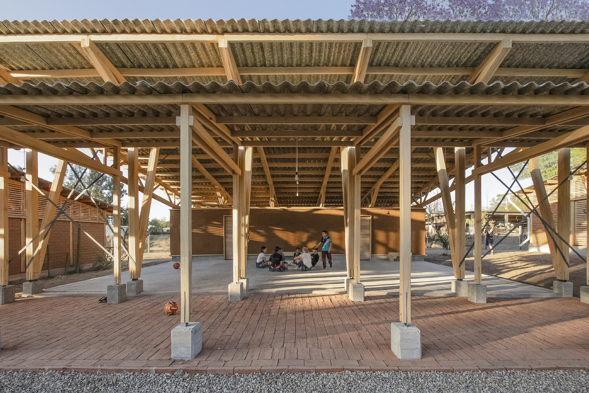 Open-air structure with diagonal timber bracing and concrete footings on brick paving, children gathered inside