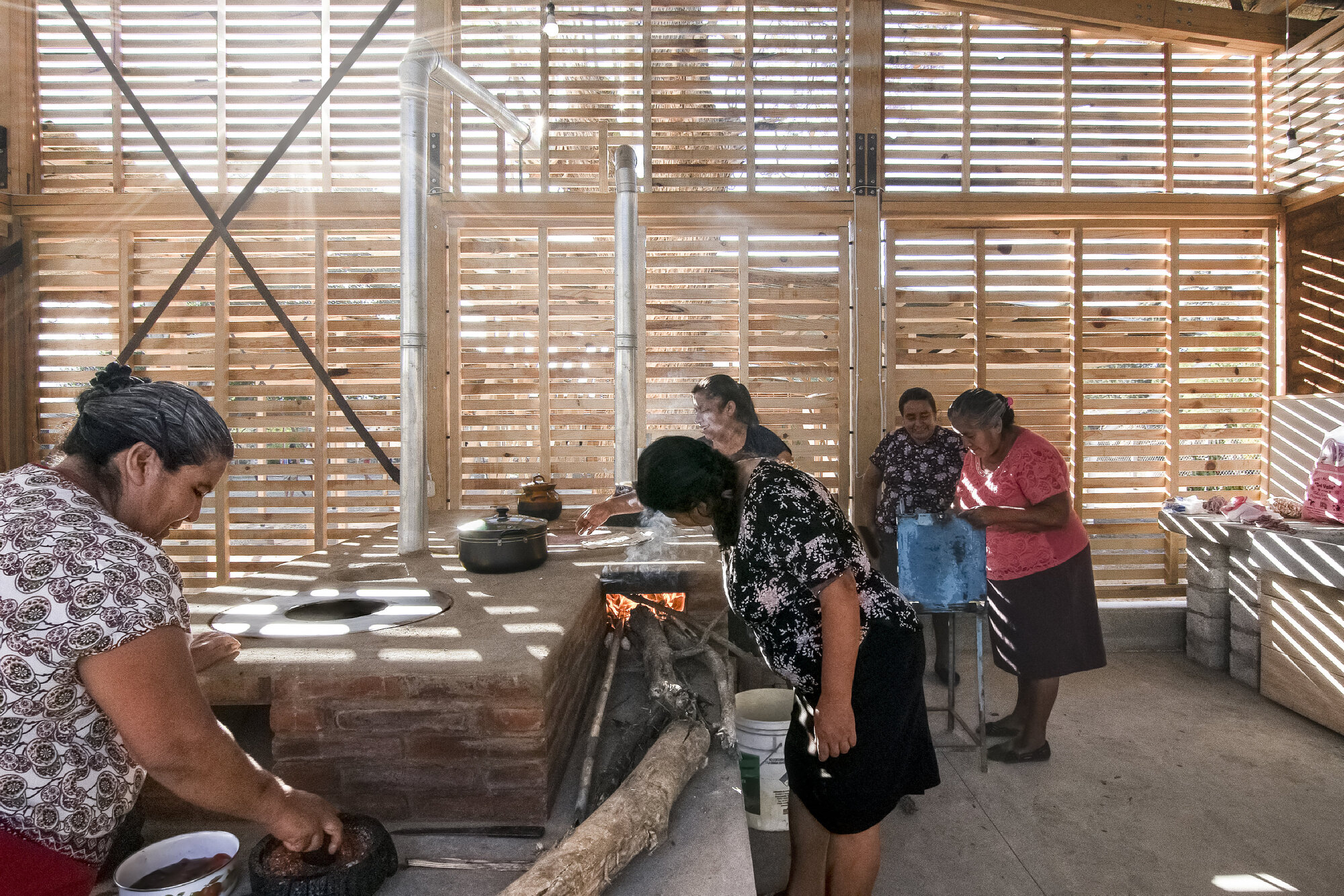 Group preparing food around the central brick hearth under exposed timber rafters and corrugated roof panels