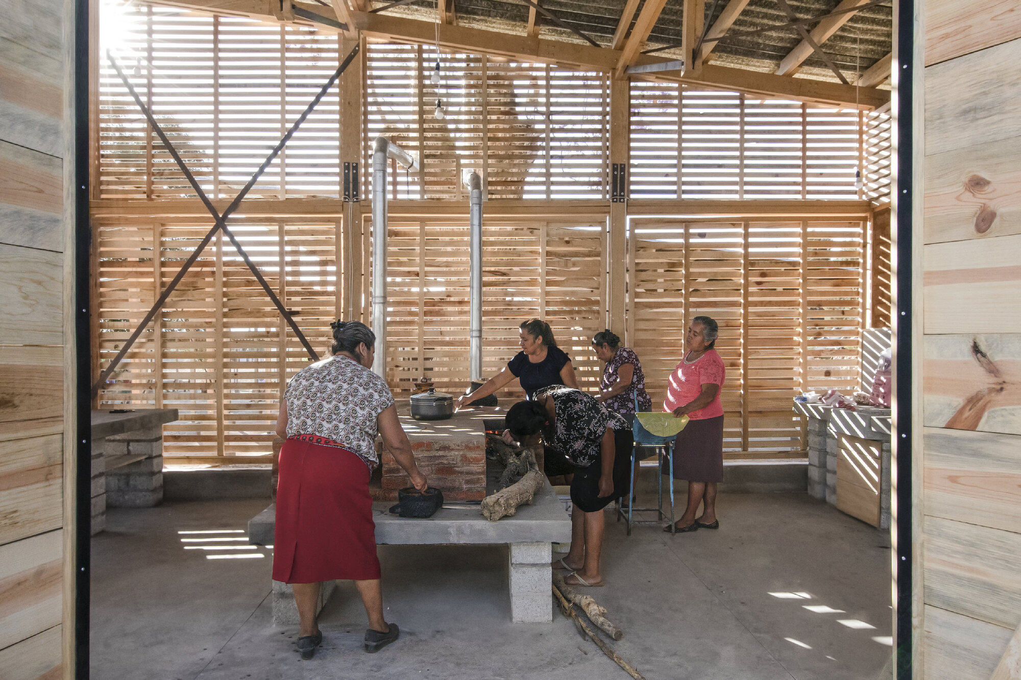 View through the steel doorframe into the cooking area with cross-braced timber walls and gathered community members