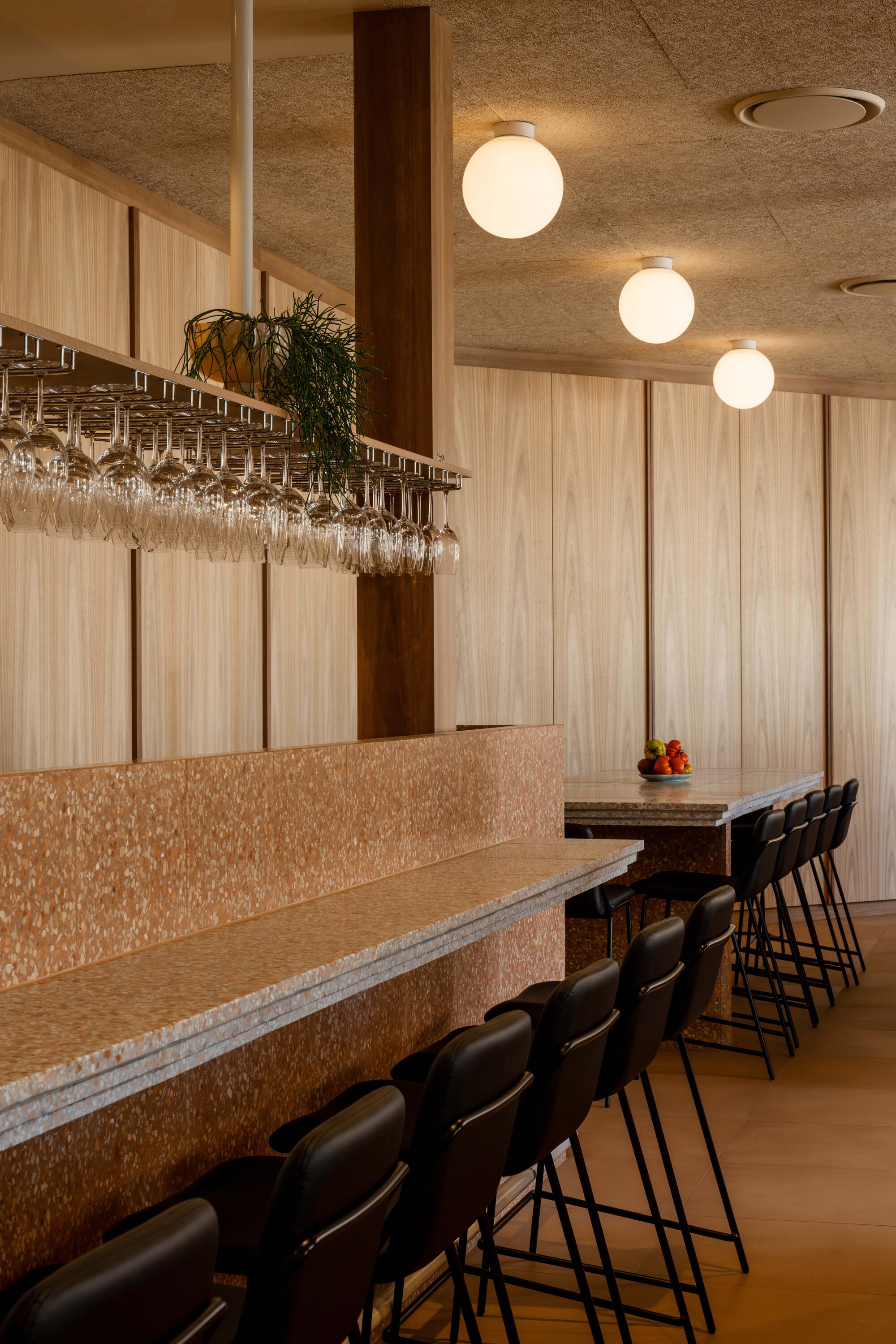 Row of bar stools along terrazzo counter with overhead wine glass rack and globe pendants