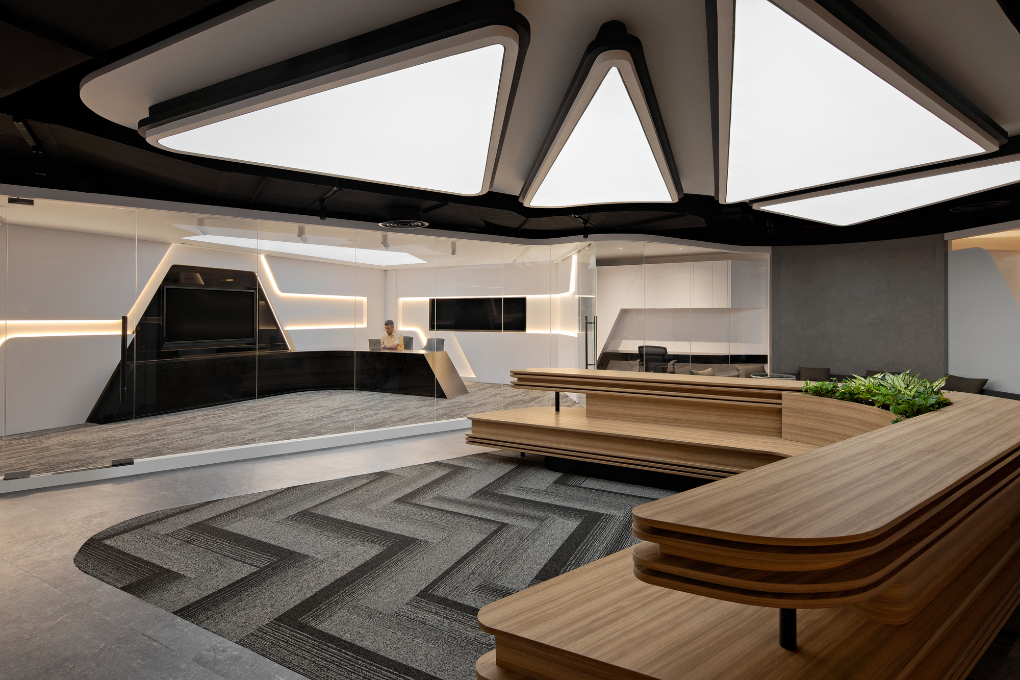 Central workspace showing layered timber desk platforms with chevron carpet patterns beneath angular ceiling luminaires