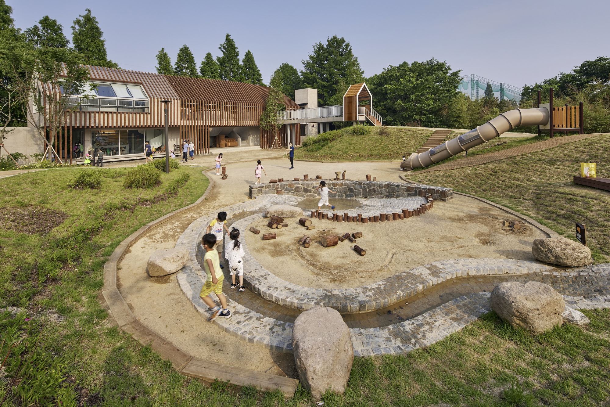Terraced sand play area with stone edging surrounded by timber-clad pavilions and children at play