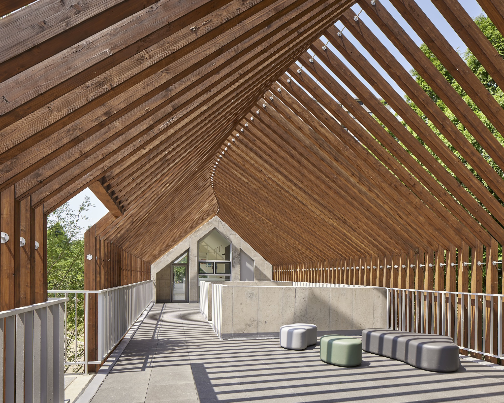 Covered outdoor terrace with slatted timber gable roof filtering afternoon sunlight onto seating pods