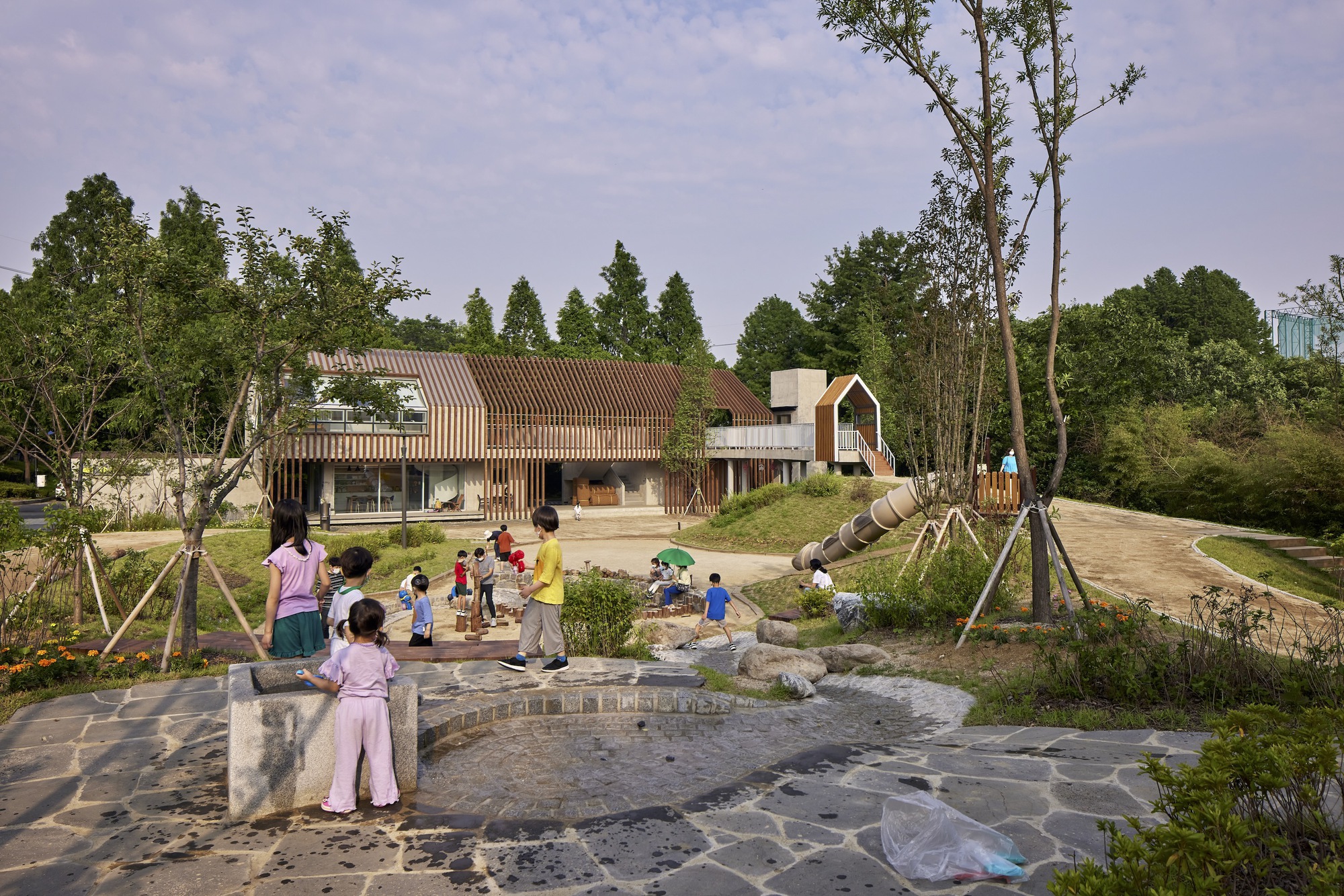 Cluster of buildings with corrugated roofs and timber screens around a landscaped courtyard where children play