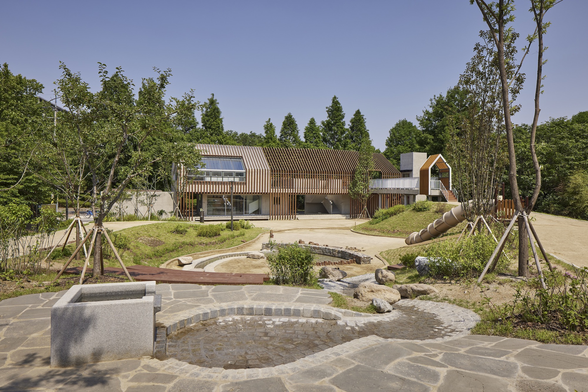 Curved stone paving and shallow water channel leading toward the timber-clad volumes under clear sky