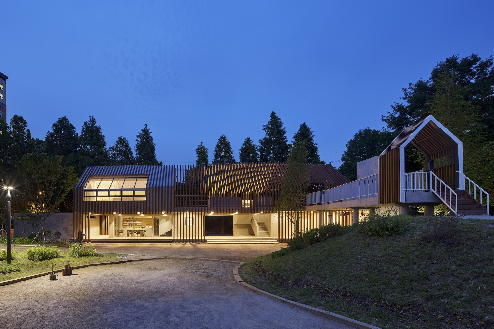 Full length view of the timber-clad volumes with illuminated interiors and sloped lawn at dusk