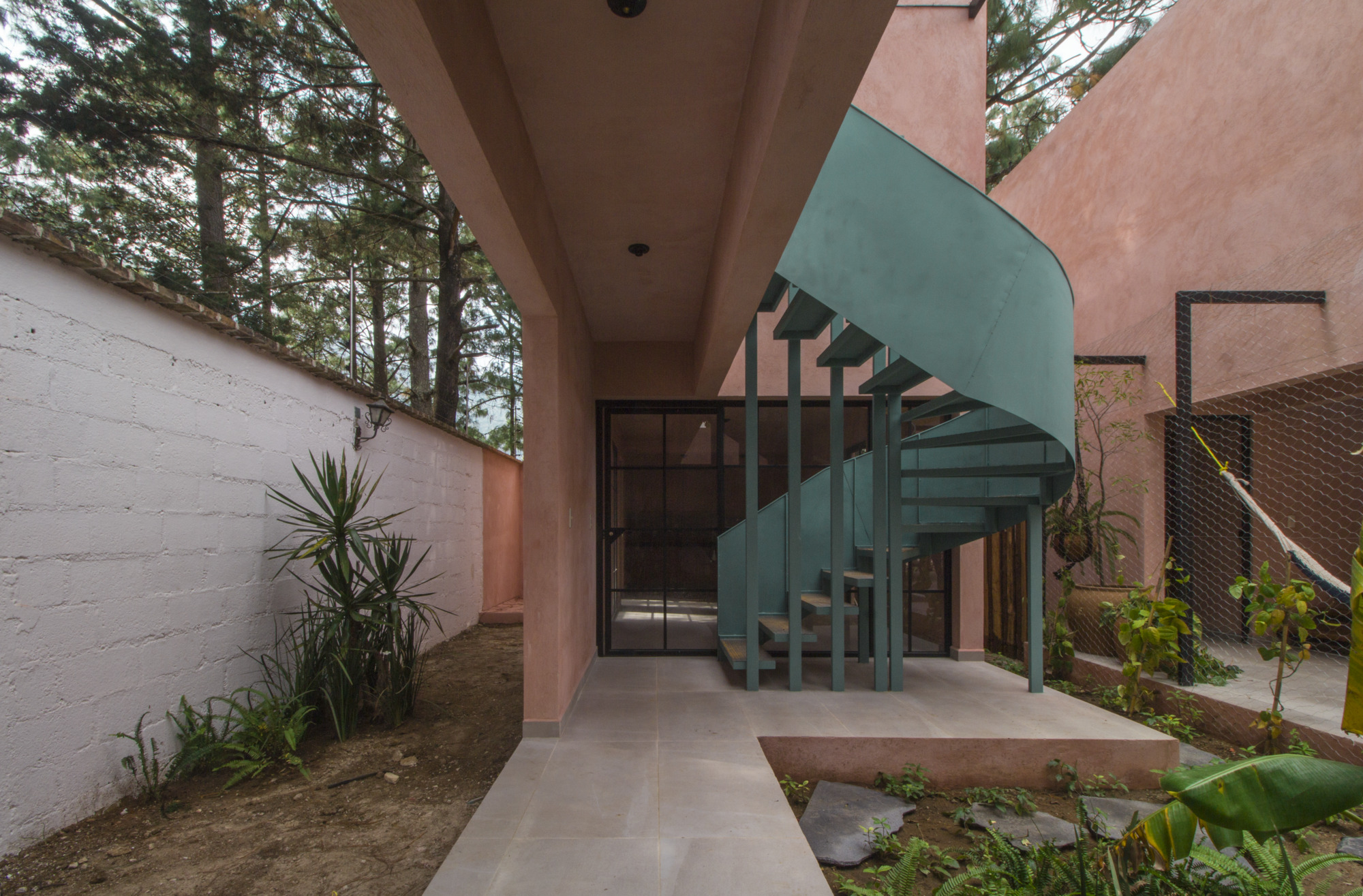 Teal spiral staircase in a courtyard with pink stucco walls and planted beds beneath pine trees