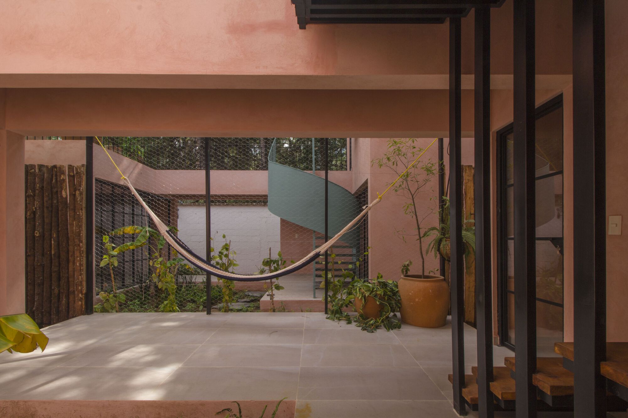 Interior view through steel-framed glazing to courtyard with hammock, spiral stair, and potted plants