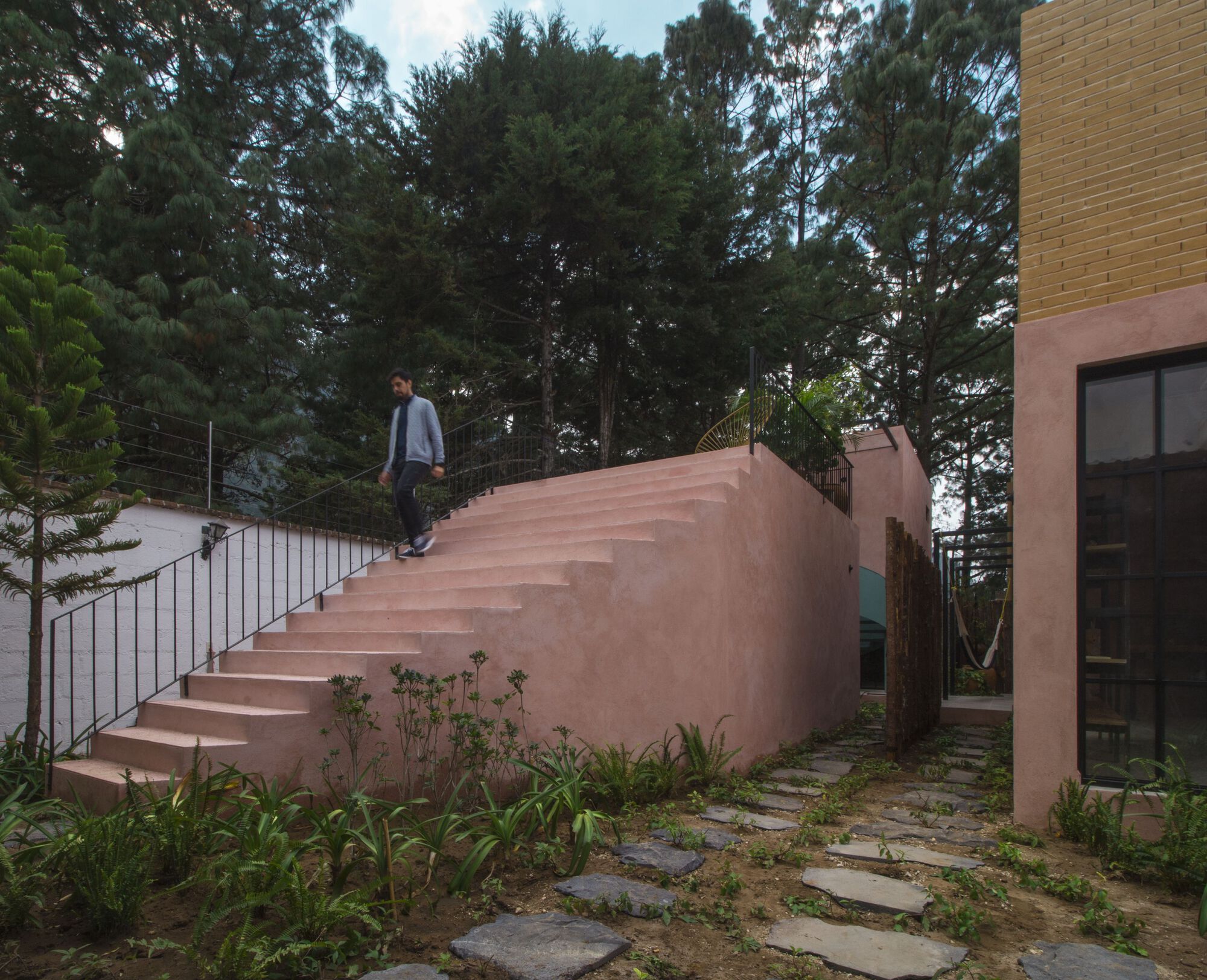 Exterior pink stucco staircase with metal railing and person ascending, surrounded by stone pavers and vegetation
