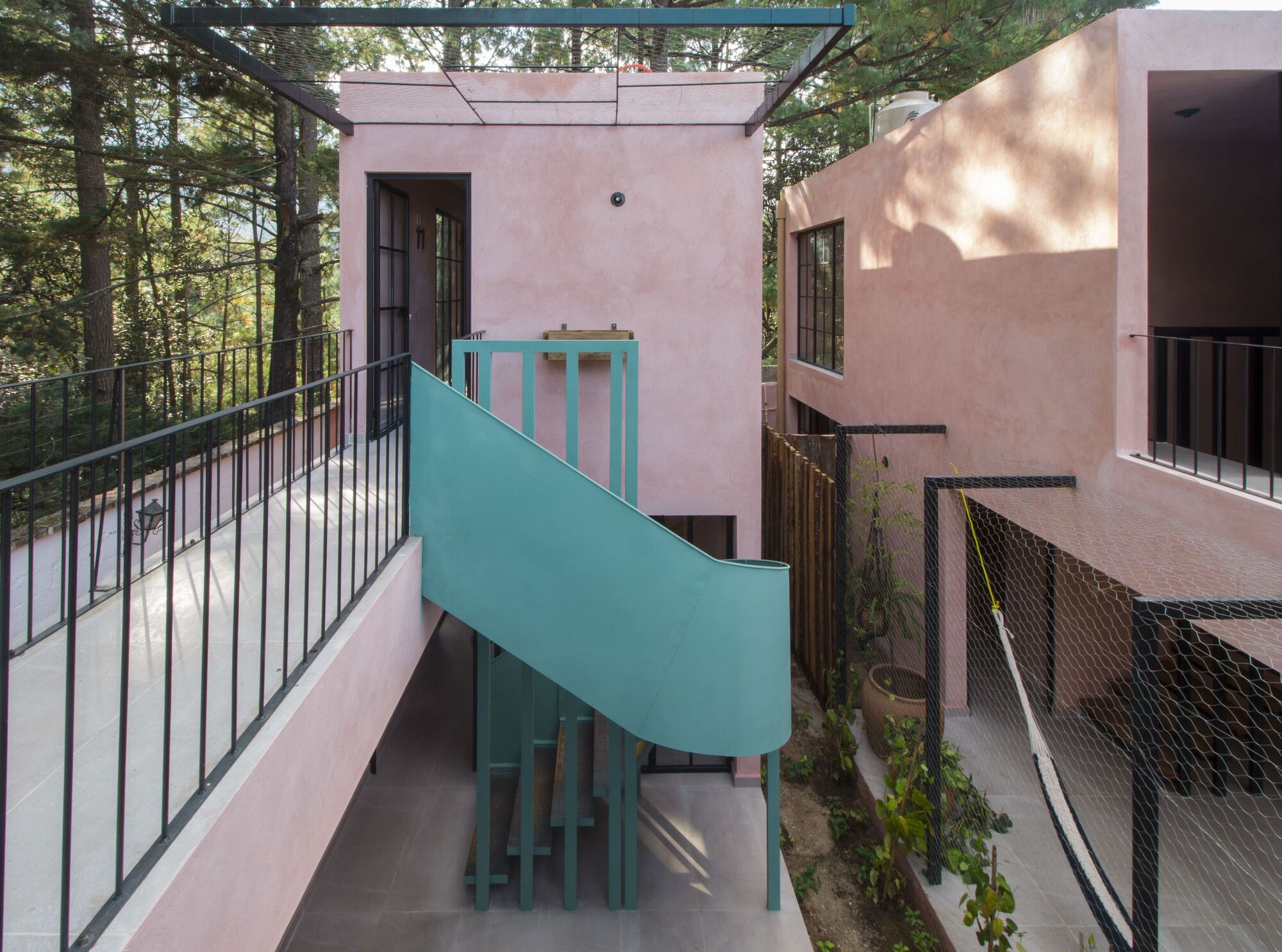 Overhead walkway with metal railing looking down at the teal spiral staircase between pink stucco volumes