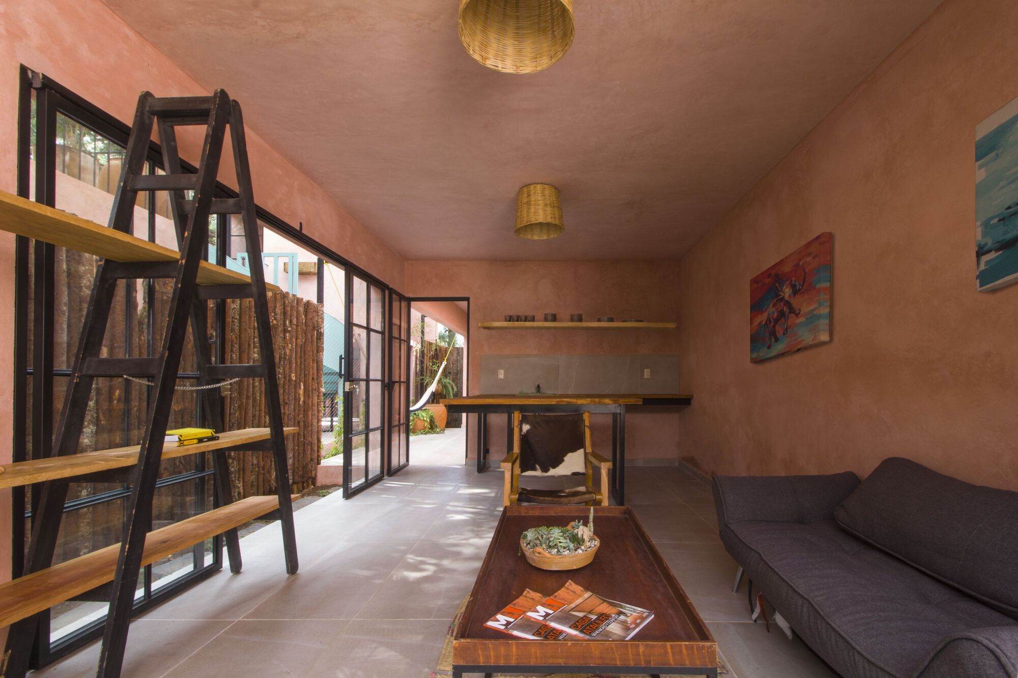 Interior living space with steel-framed glazing, ladder shelving, woven pendant lights, and pink stucco walls