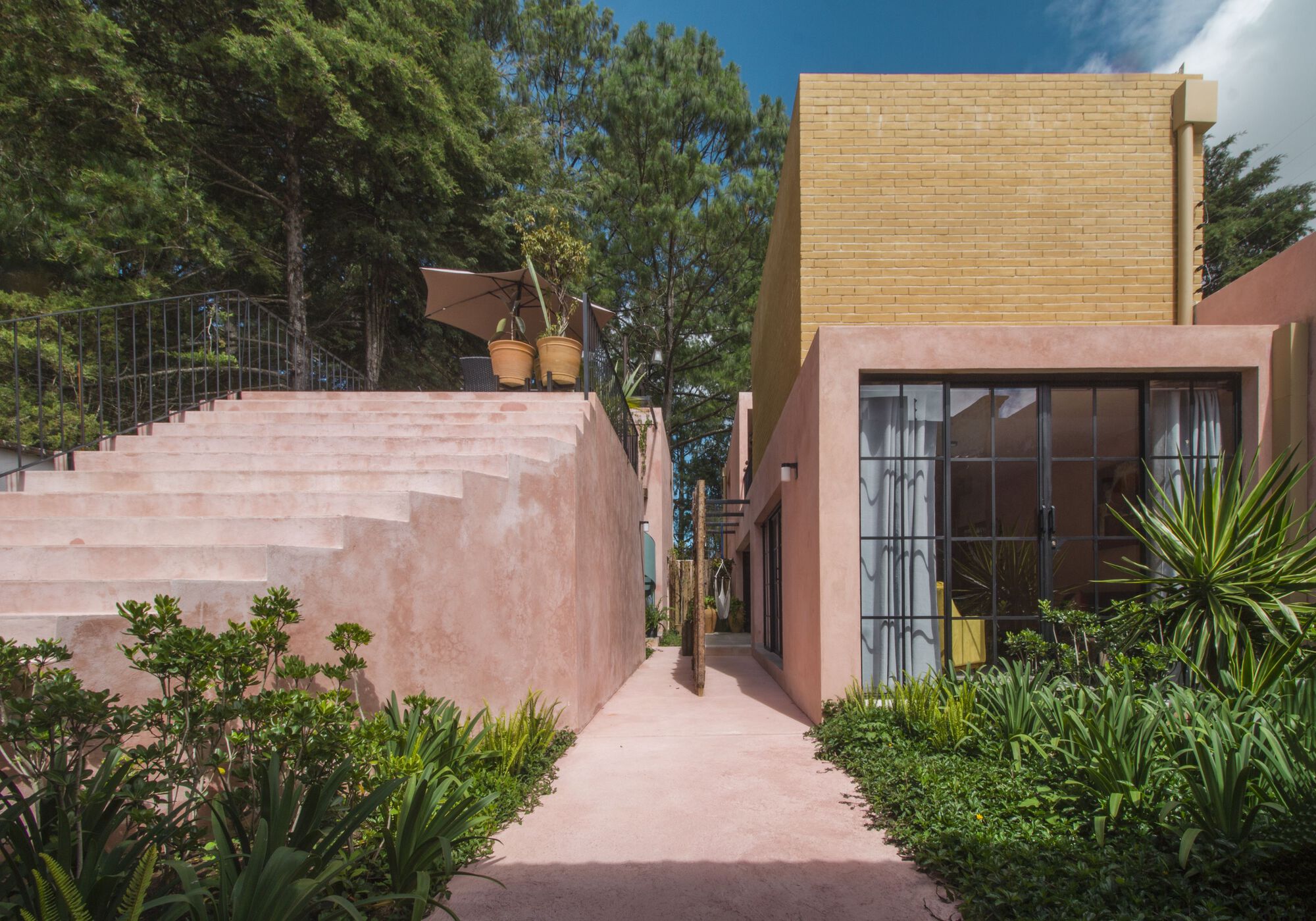 Pink stucco pathway winding past gridded glazing and a yellow brick volume among tropical planting