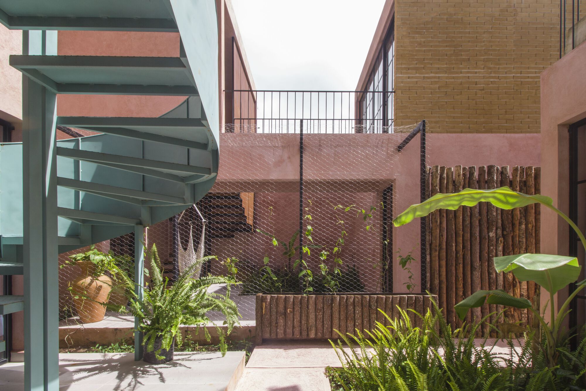 Courtyard with pink textured walls, vertical timber screening, and potted tropical plants beneath an overcast sky