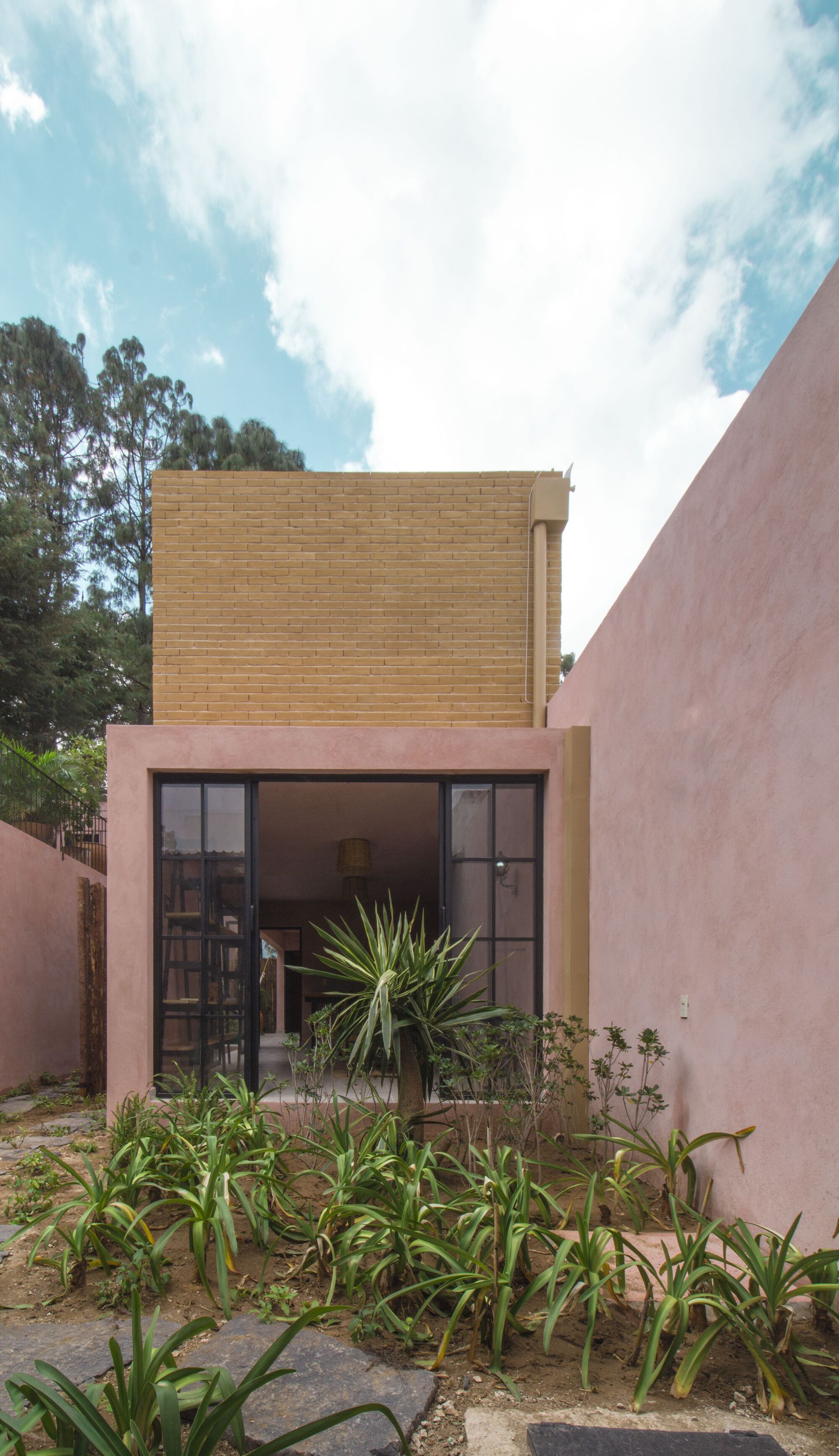 Garden facade with black-framed glazed door beneath a tan brick volume and lush planted bed