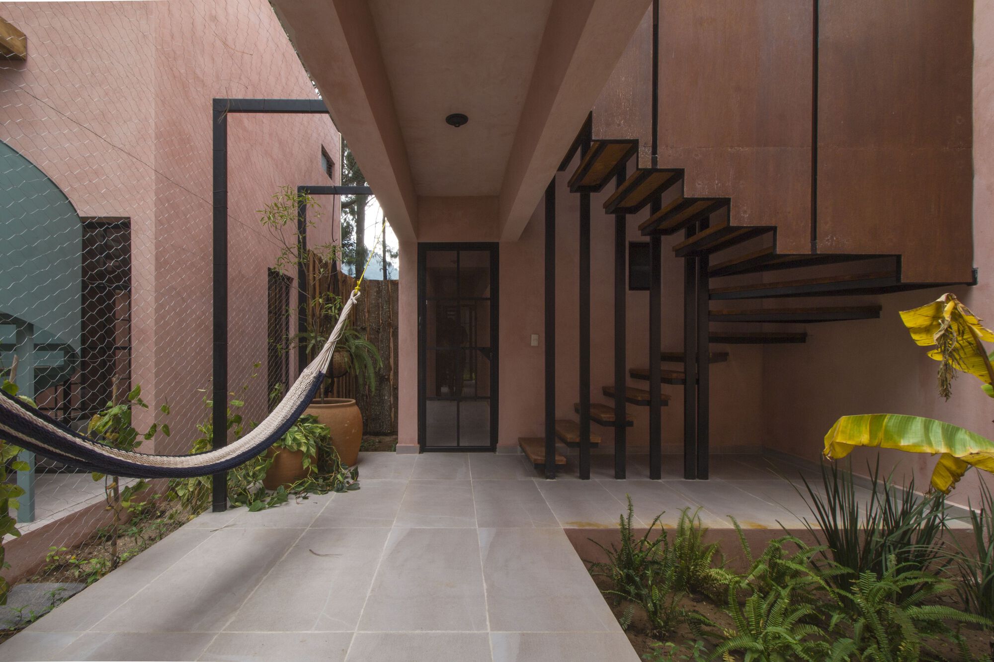 Tiled passageway with steel-framed staircase, hammock, and tropical plants against pink stucco walls