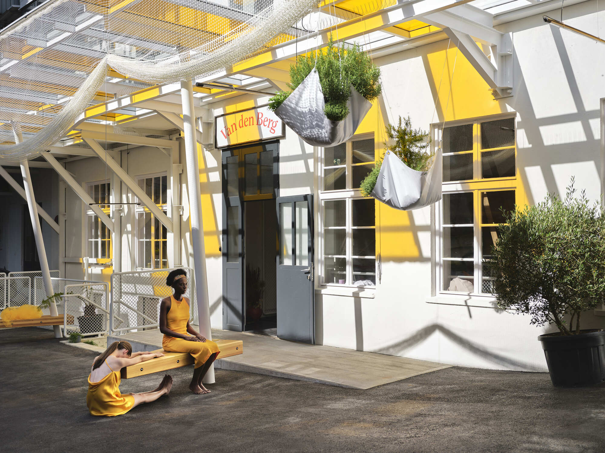 Courtyard with white tubular canopy structure, yellow-framed windows, and two figures seated on a bench in sunlight