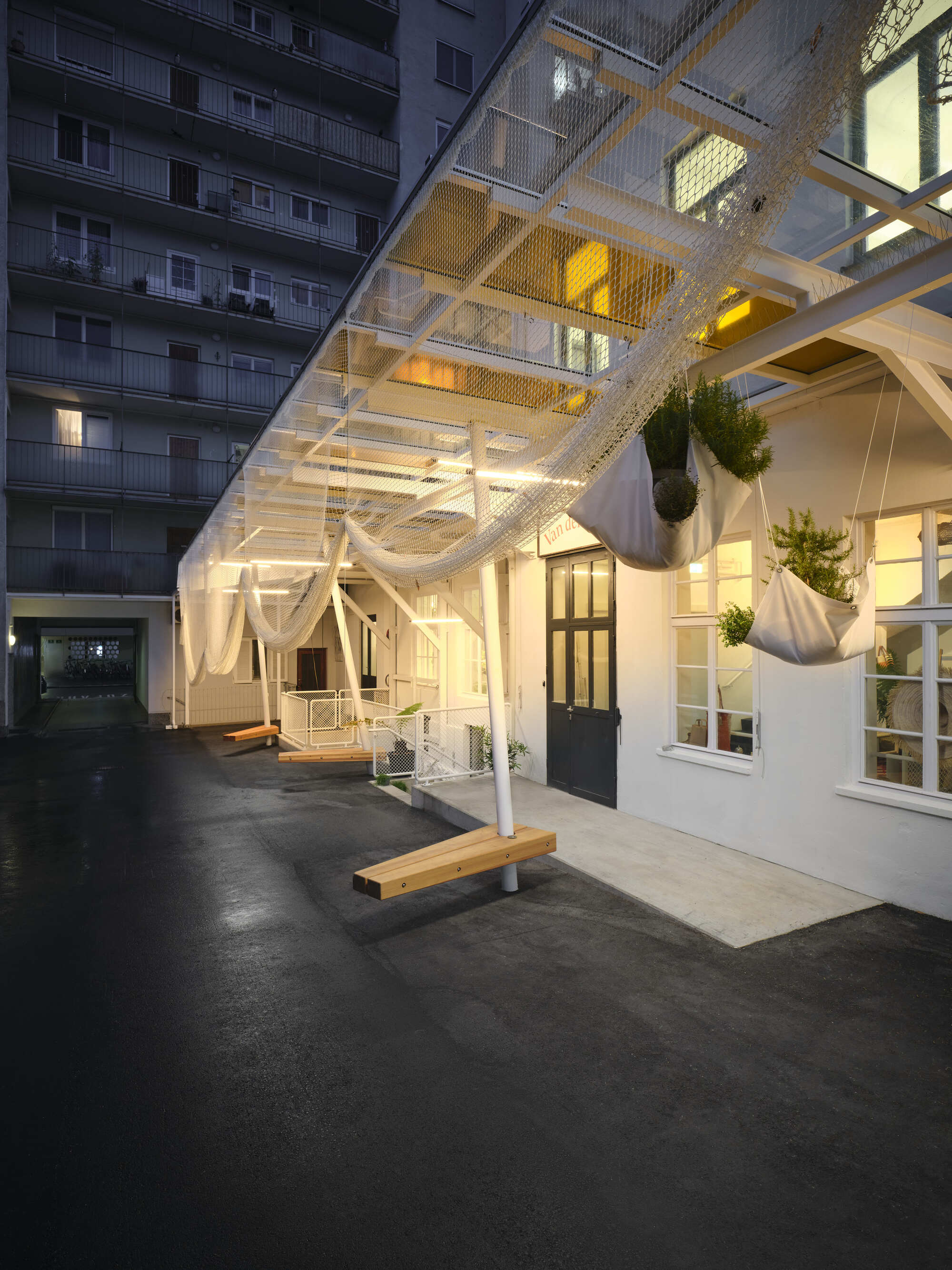 Courtyard at dusk showing illuminated white metal canopy structure with hanging planters and adjacent apartment tower
