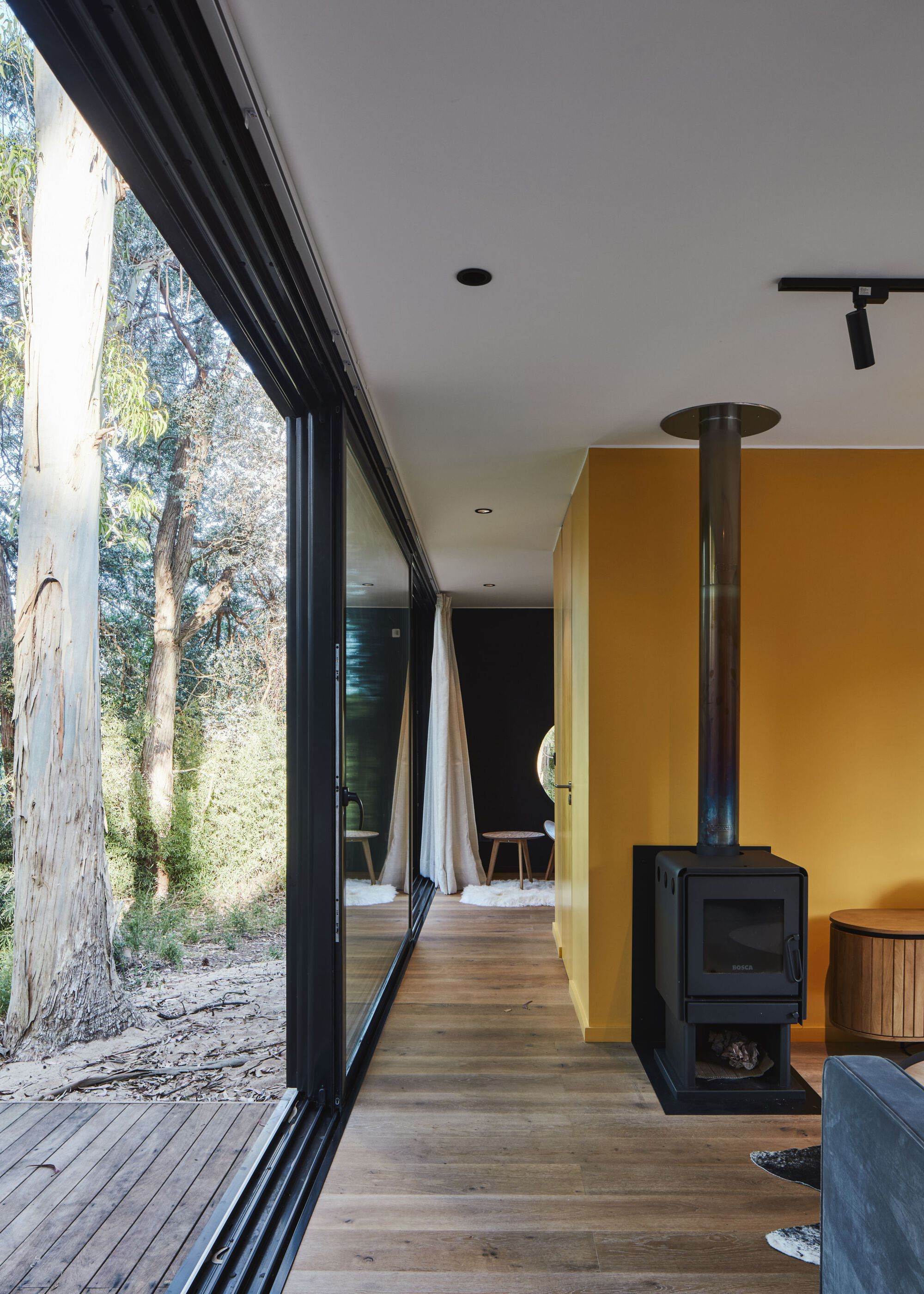 Interior corridor with freestanding black wood stove against a yellow wall and floor-to-ceiling glazing overlooking trees