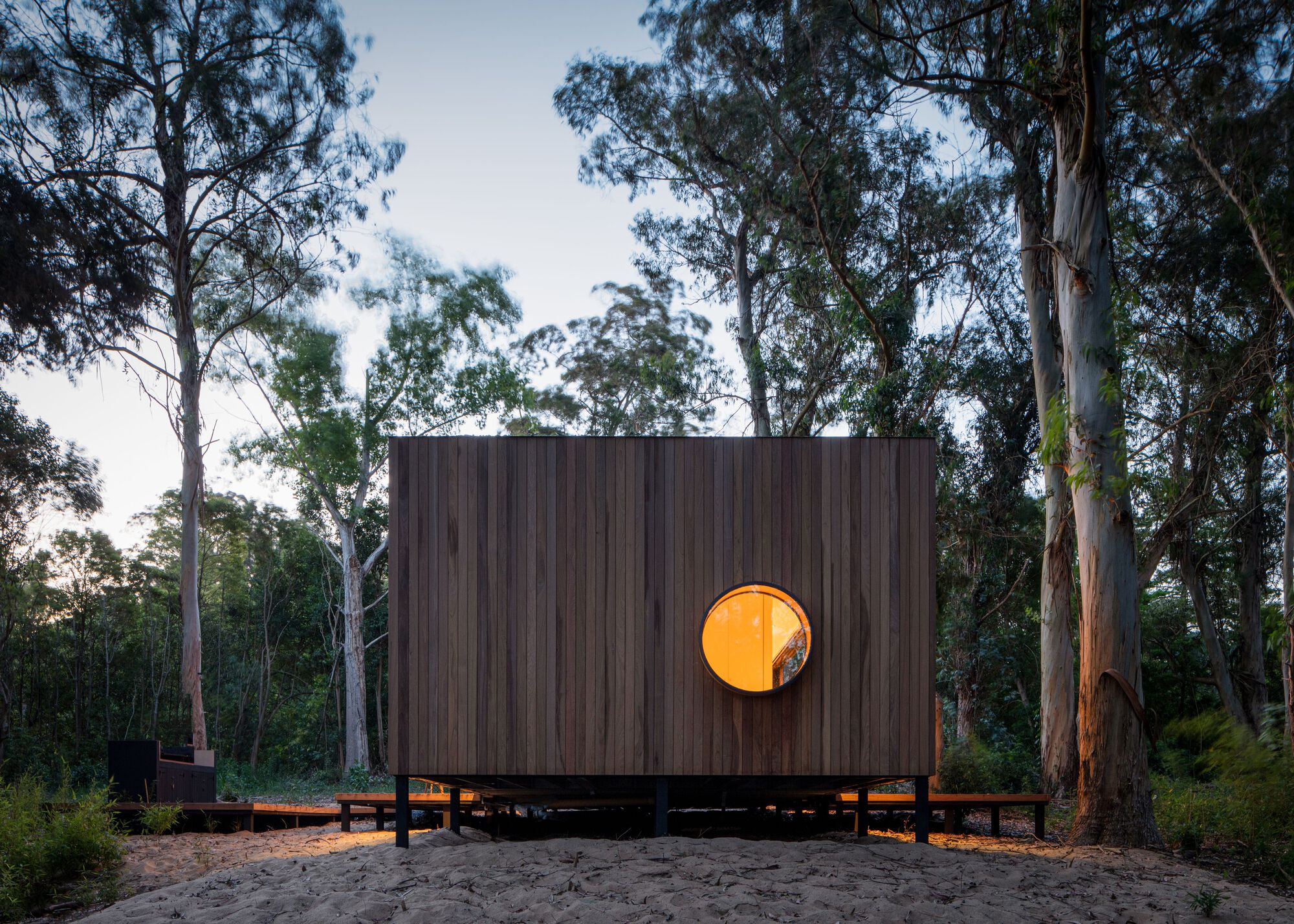 Rear facade with illuminated circular window piercing the vertical timber cladding at dusk