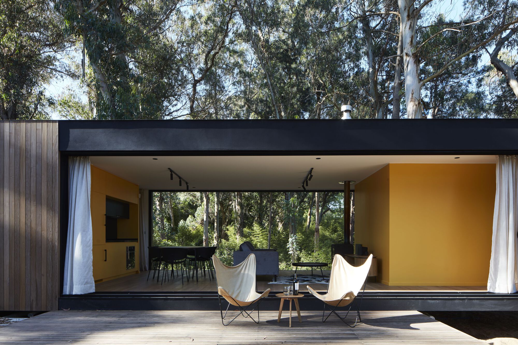 Covered terrace with butterfly chairs on timber decking beneath a flat black soffit looking into the living space