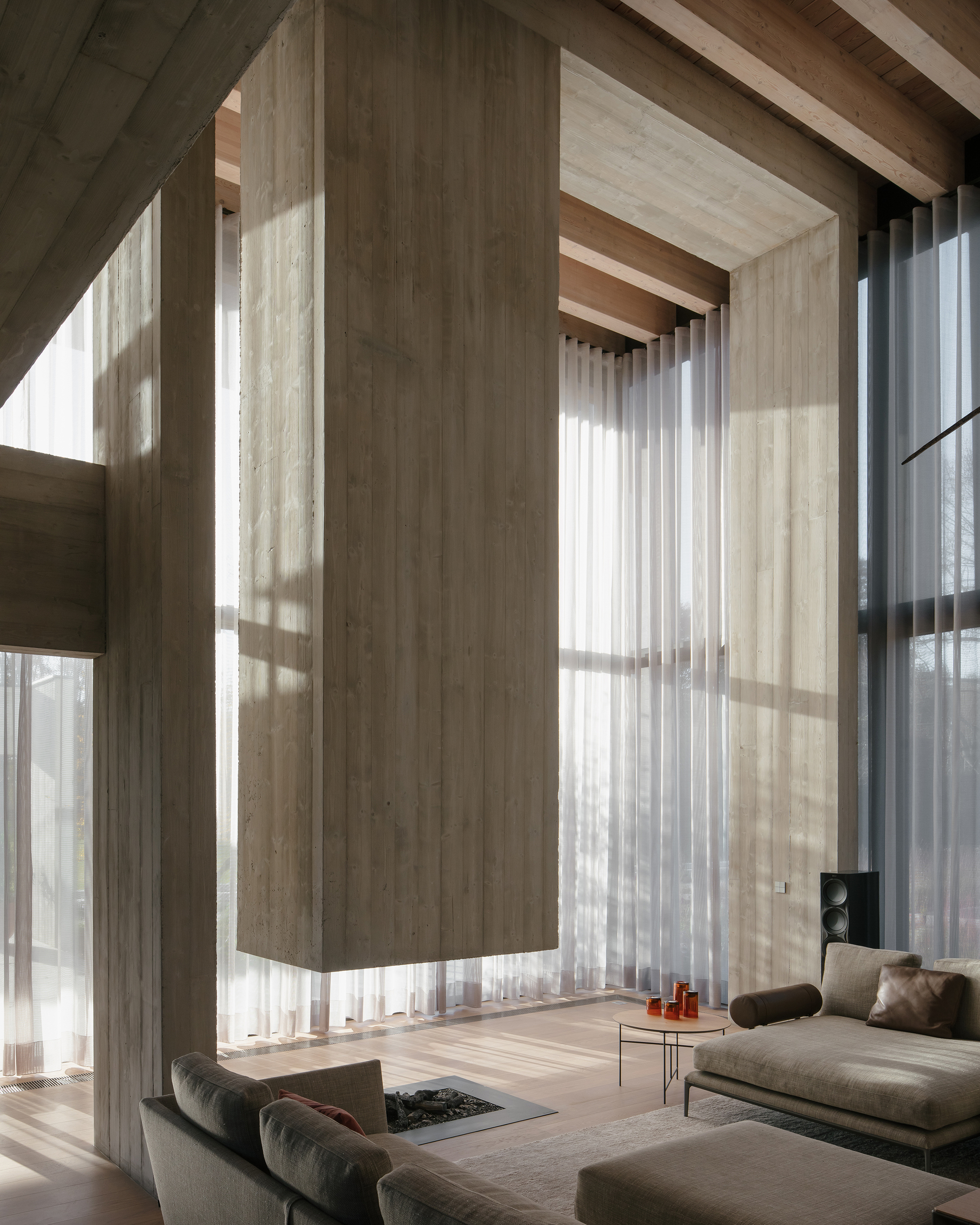 Double-height living space with floor-to-ceiling windows, timber ceiling beams and sheer curtains filtering sunlight