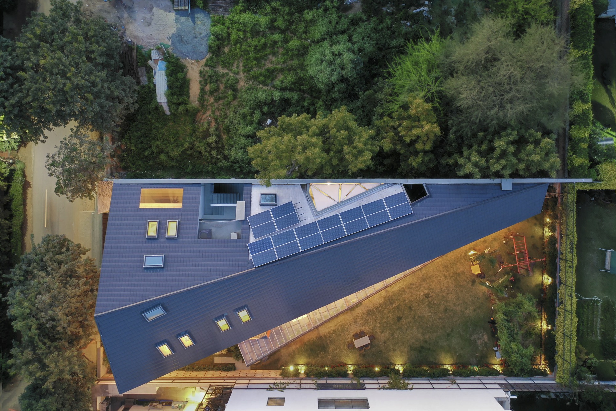 Aerial view of the angular roof with solar panels and skylights surrounded by dense vegetation at dusk
