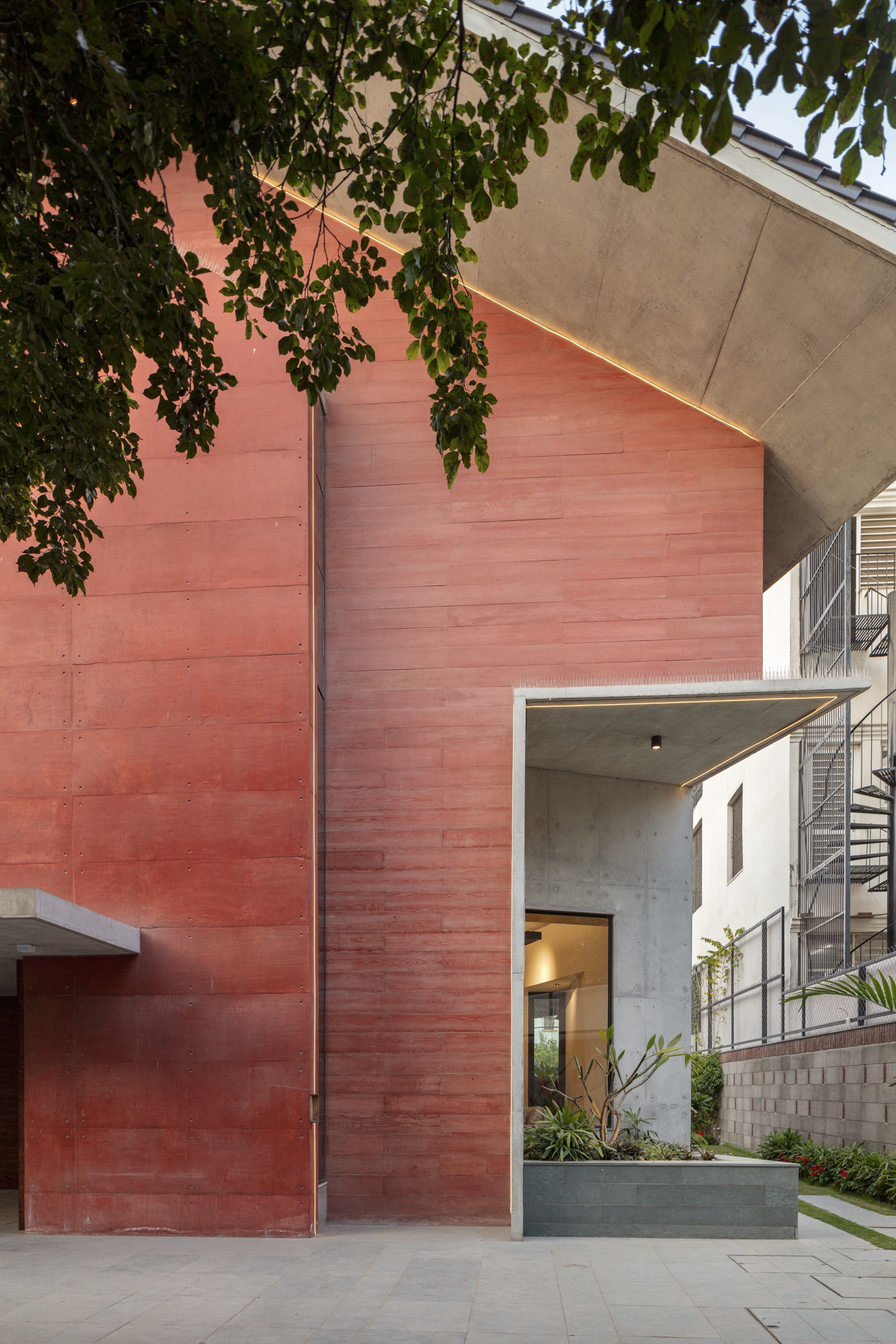 Red stone wall surface meeting concrete entrance volume framed by overhanging tree branches at dusk