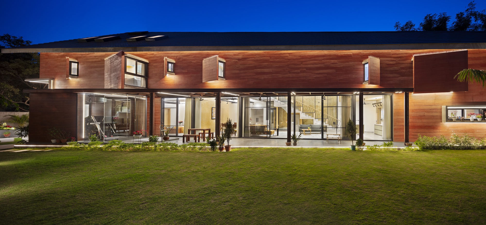 Rear elevation with horizontal timber cladding and continuous glazed openings illuminated at twilight above a lawn