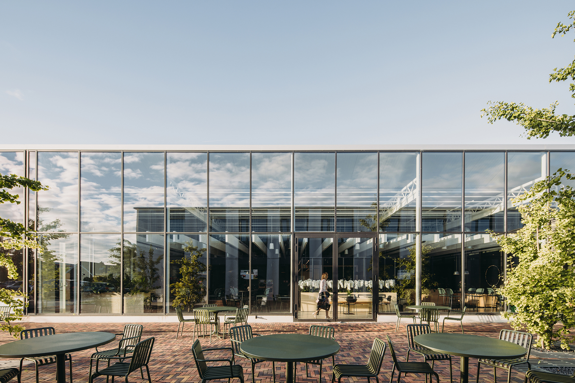 Floor-to-ceiling glass facade reflecting clouds with outdoor brick terrace and metal cafe tables