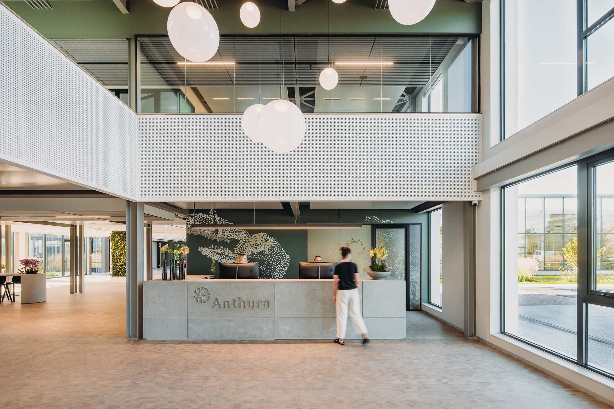 Double-height reception lobby with concrete desk, spherical pendant lights, and a person walking past glazed walls