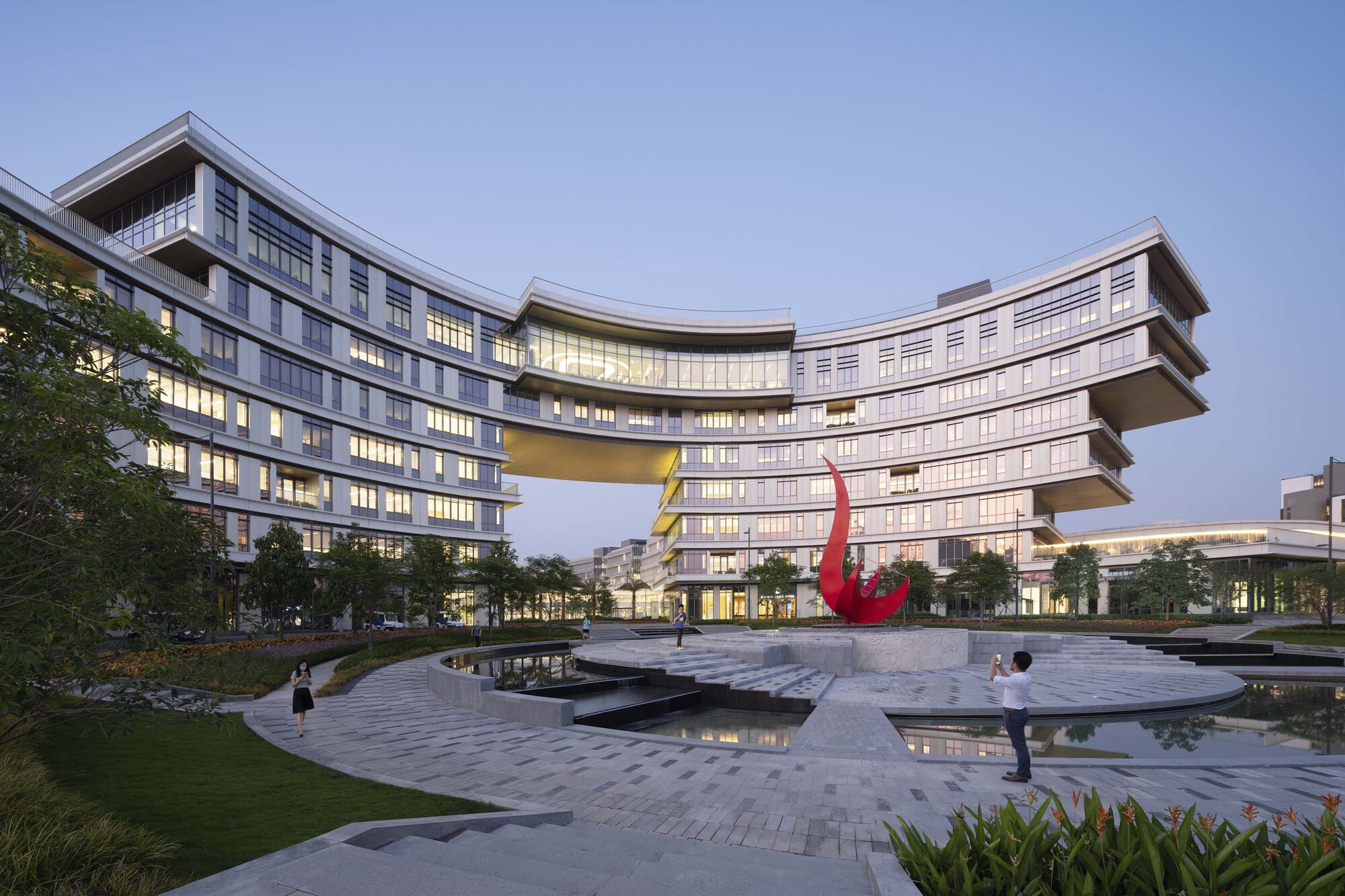 Courtyard with reflecting pools and red sculpture beneath a curving skybridge connecting two multi-story wings at dusk
