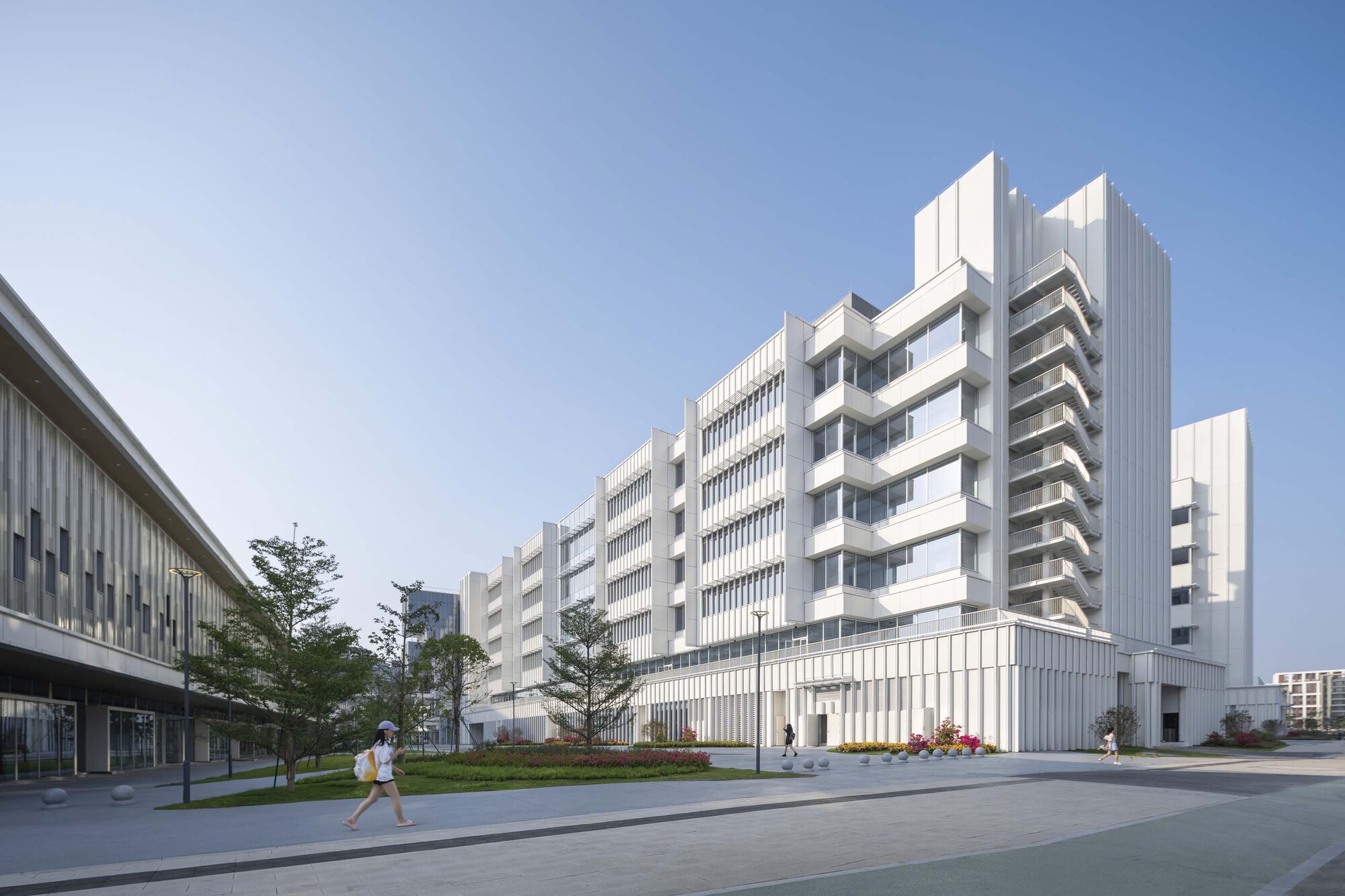 White vertically ribbed facade with stepped massing and a lone pedestrian walking along the tree-lined plaza