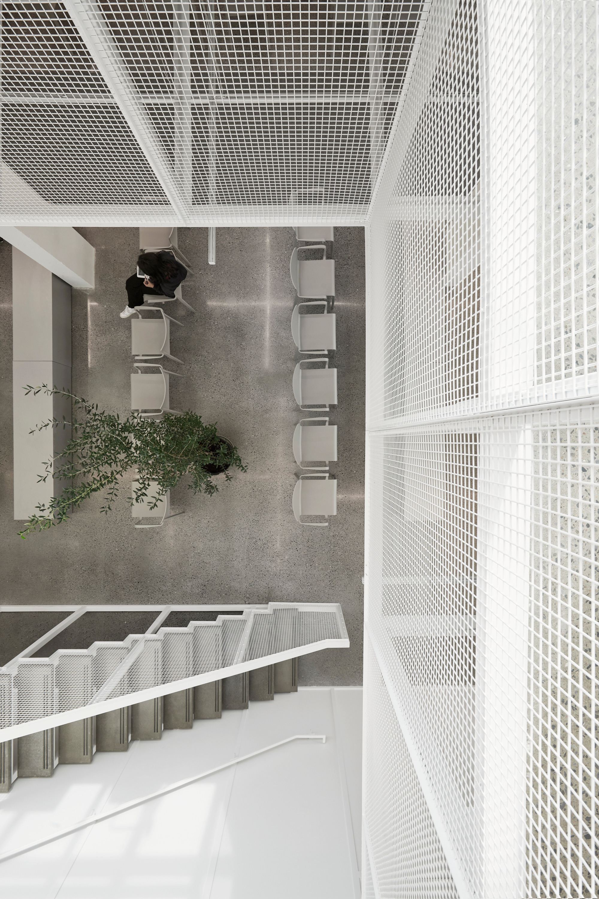 Top-down view of a multi-story atrium with white perforated metal balustrades and a person seated at grey tables