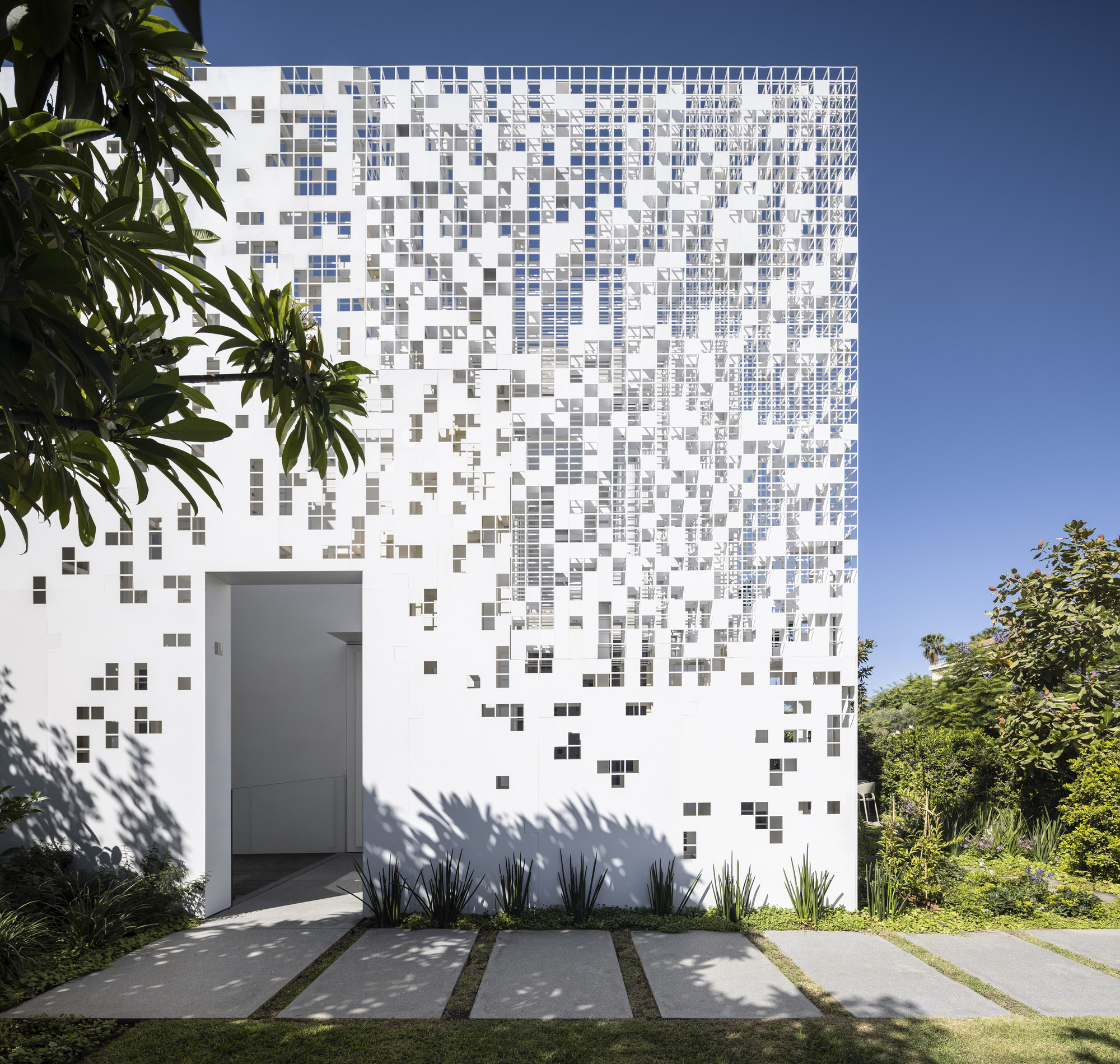 White facade with gradient pixelated tile pattern framed by garden plants and concrete pavers