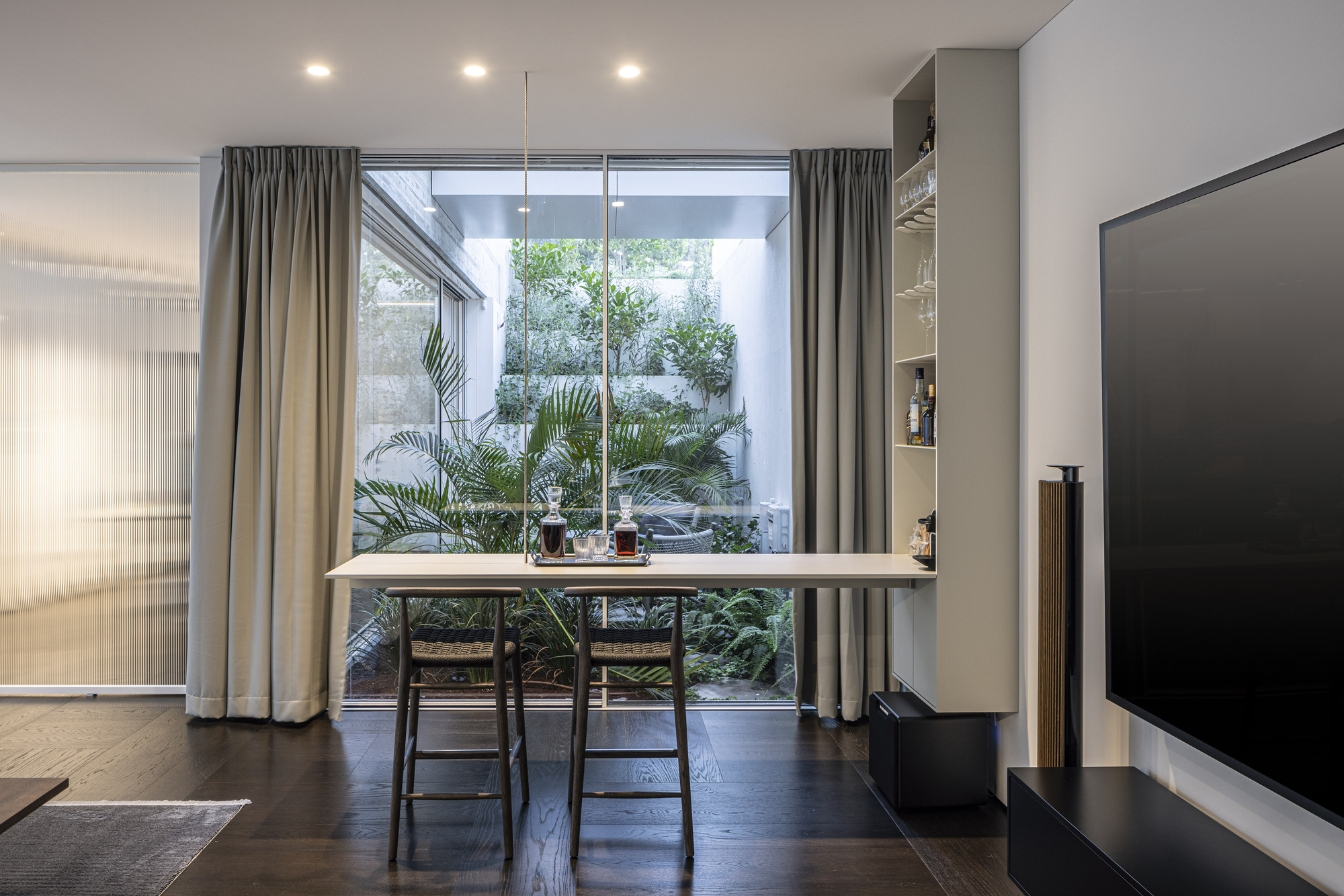 Dining area with two chairs facing a glazed courtyard filled with potted palms and greenery