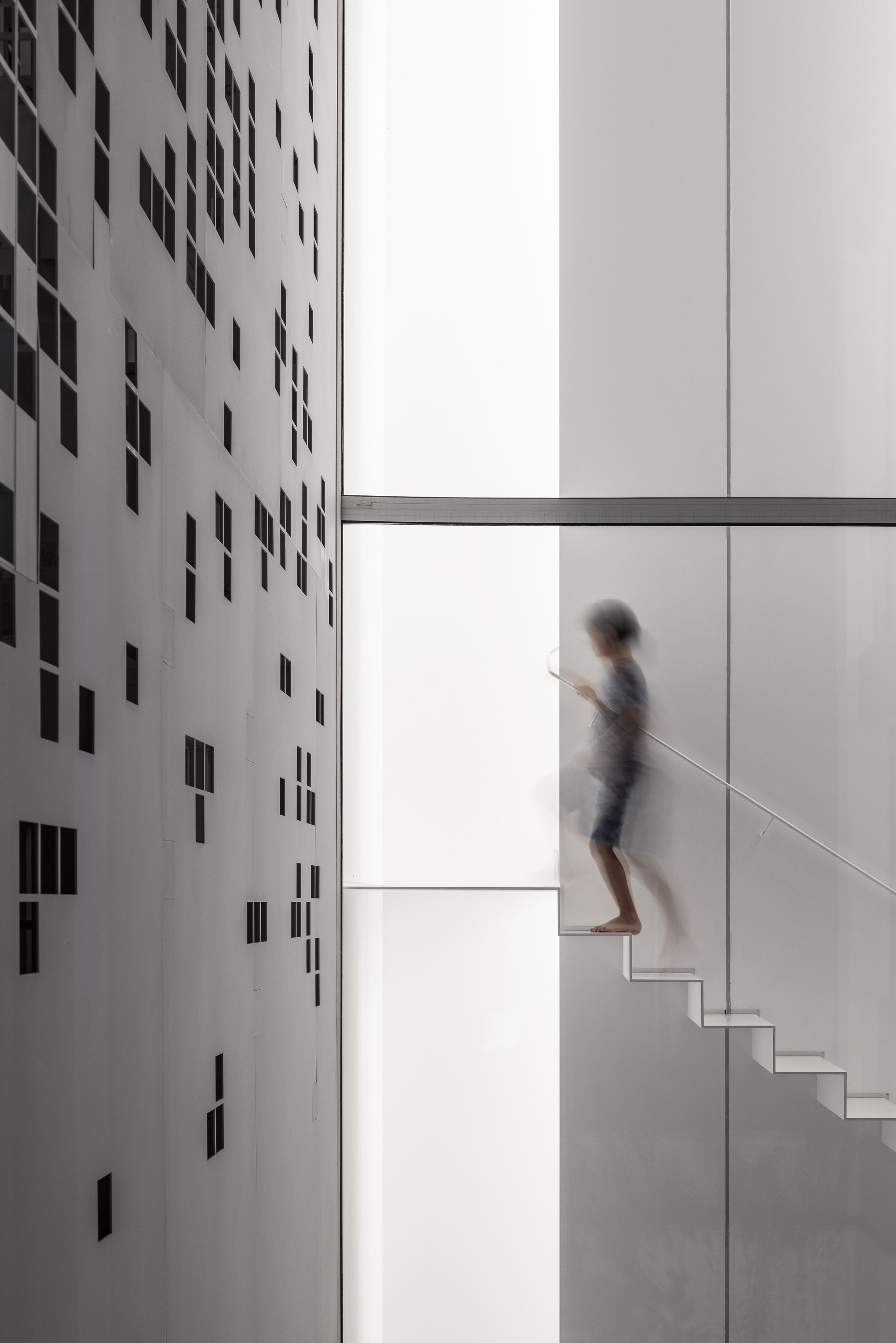 Person ascending a white staircase beside a perforated wall casting scattered shadows in bright daylight