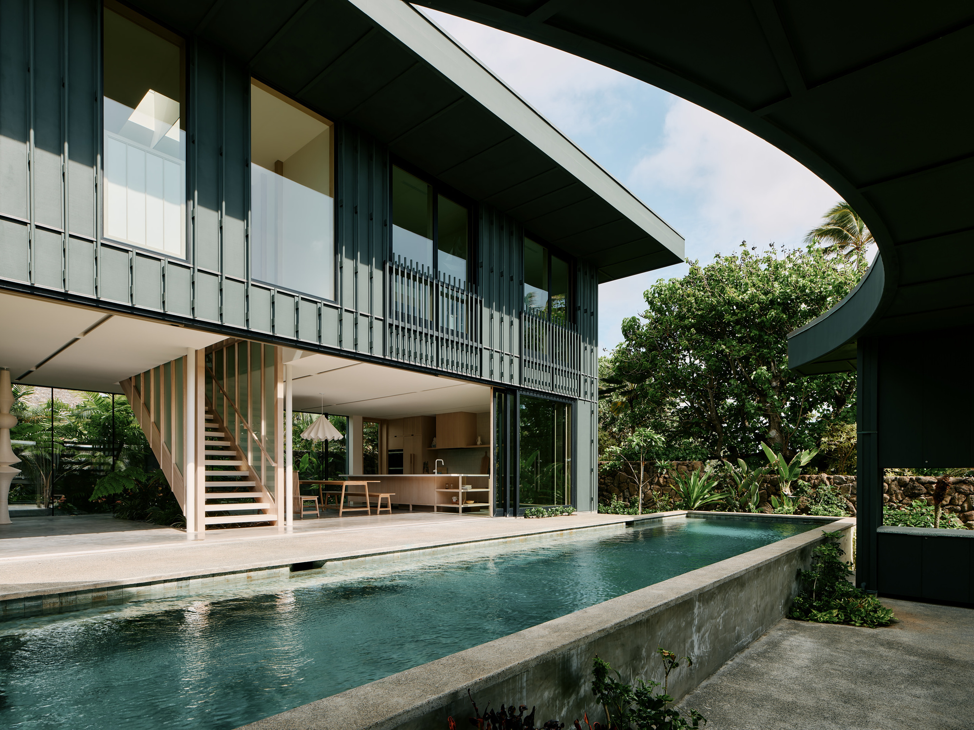 Elevated lap pool with concrete walls beside an open-plan living area and timber stair under a curved roof