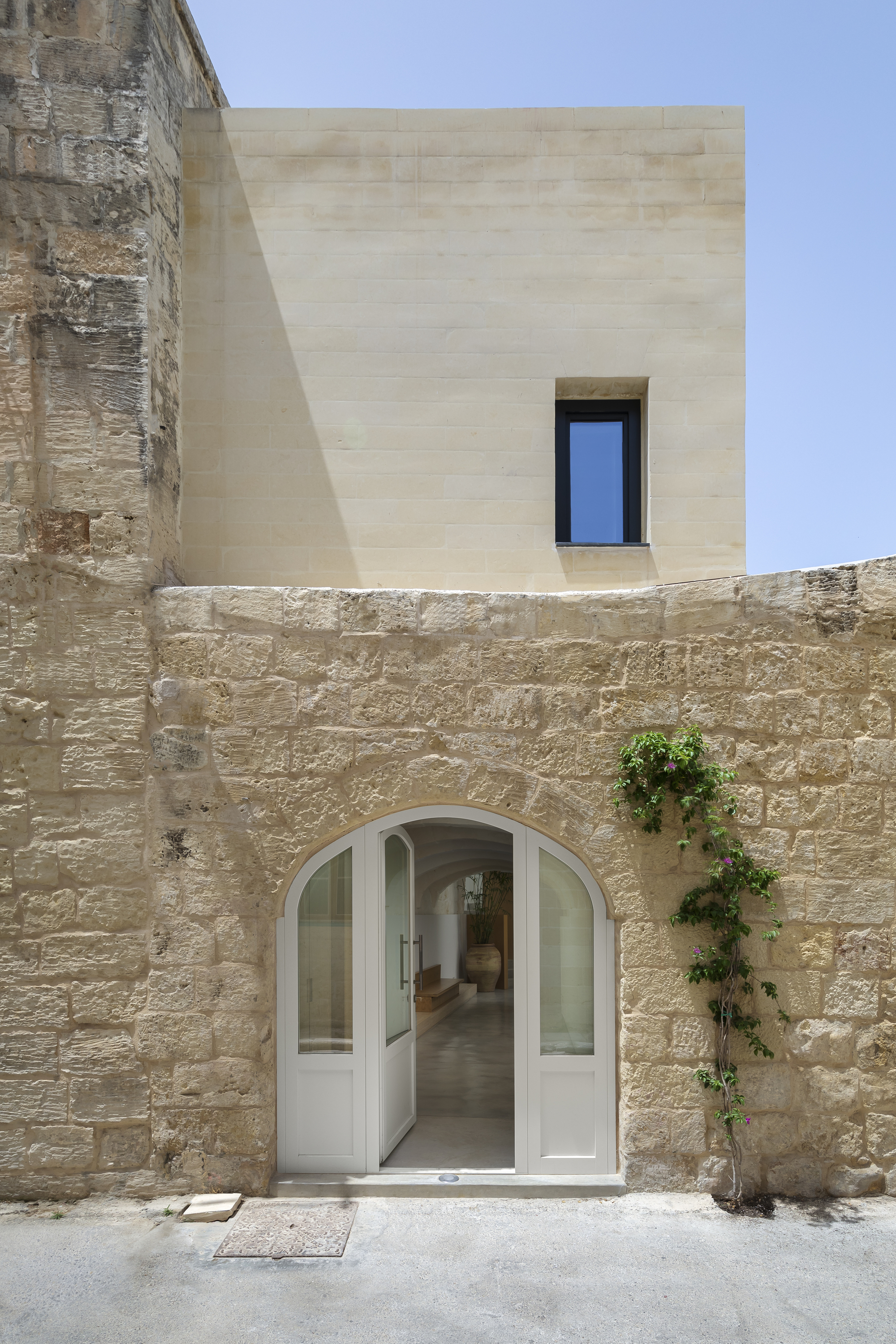 White arched doorway set into weathered limestone wall below a smooth rendered upper storey with single window